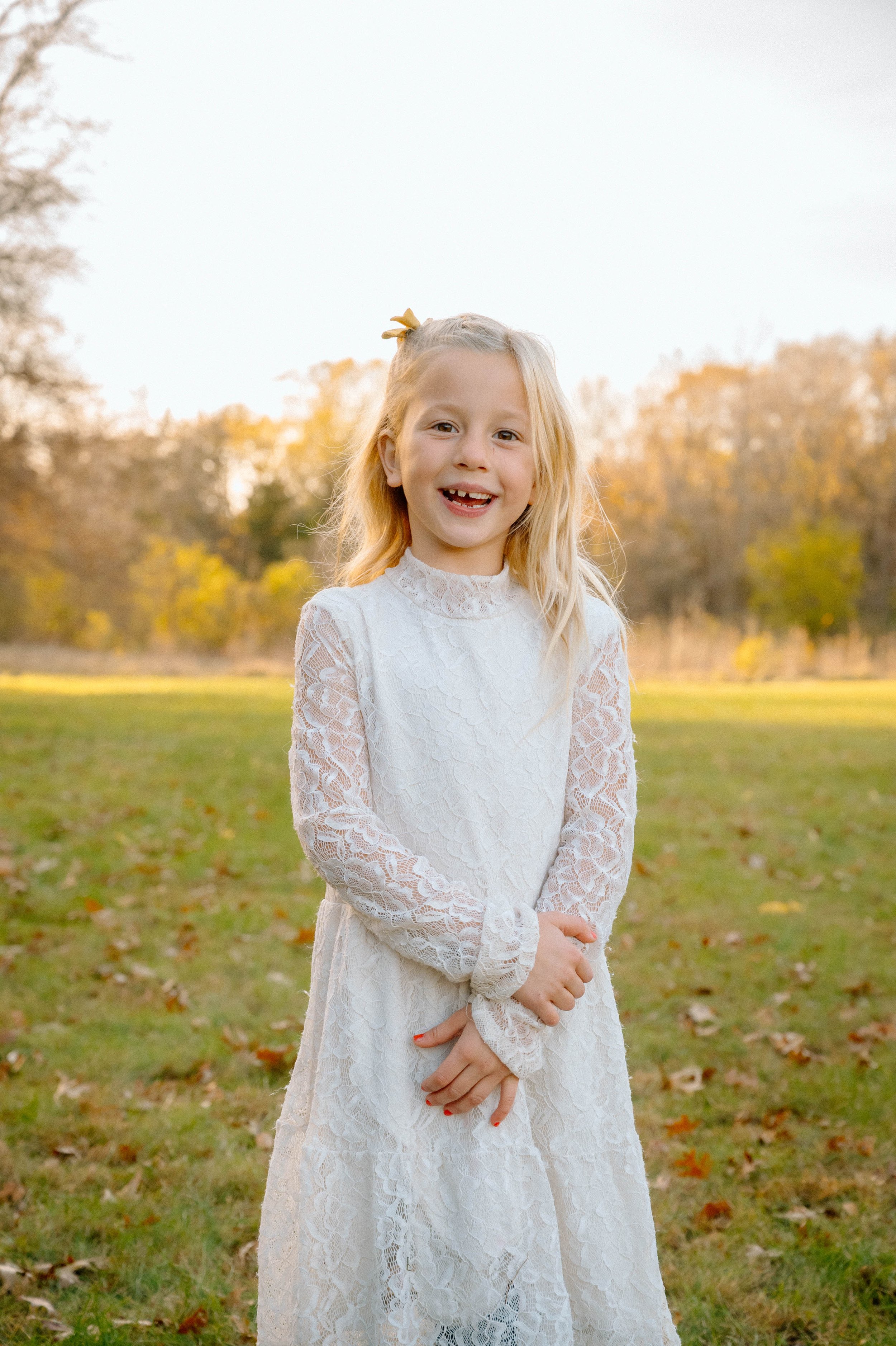 A young girl with blonde hair wearing a white lace dress, standing outdoors on a grassy field during autumn, smiling at the camera.