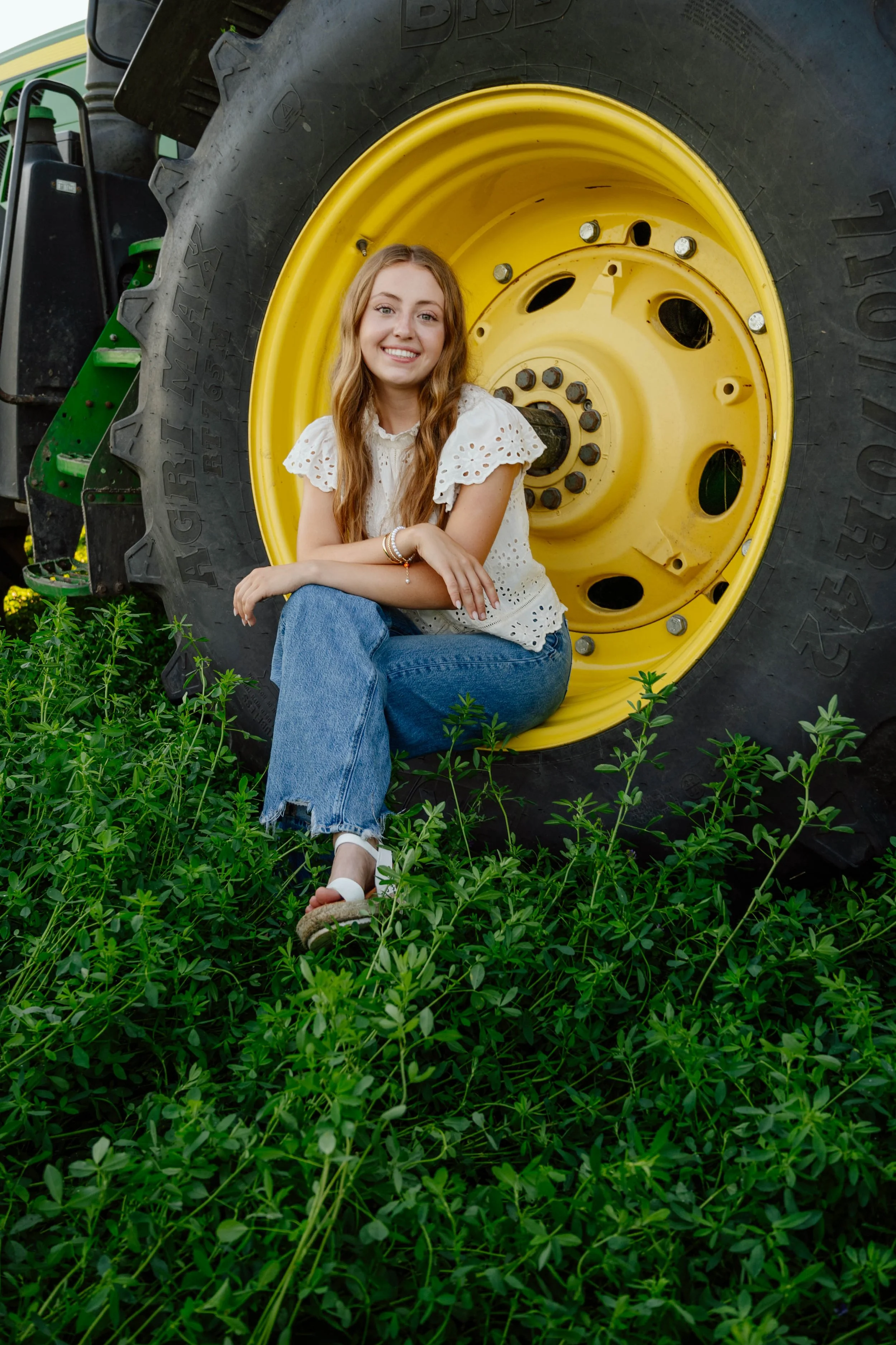 A young woman with long, wavy hair wearing a white eyelet blouse and blue jeans, sitting on the ground among green plants, leaning against a large yellow tractor tire, smiling at the camera.