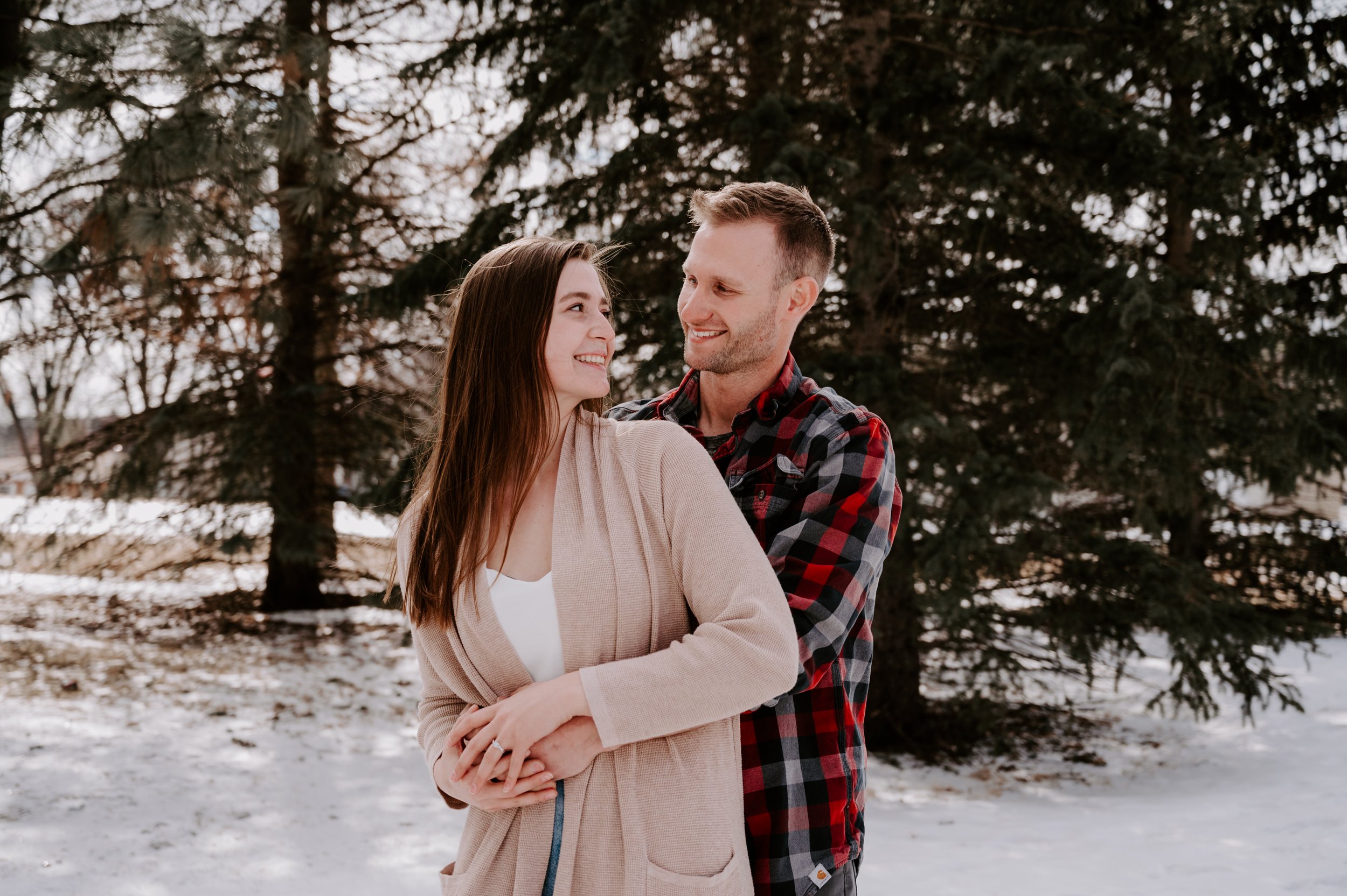 A couple standing close together outdoors in a snowy landscape with tall evergreen trees, smiling at each other.
