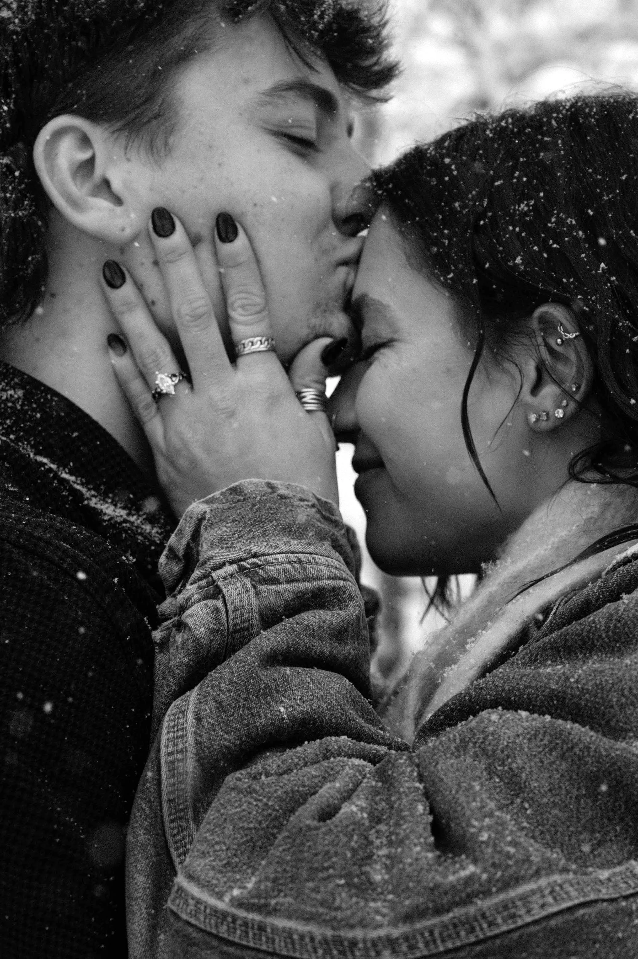 A black and white close-up of two people sharing a kiss, with snow on their hair and shoulders.