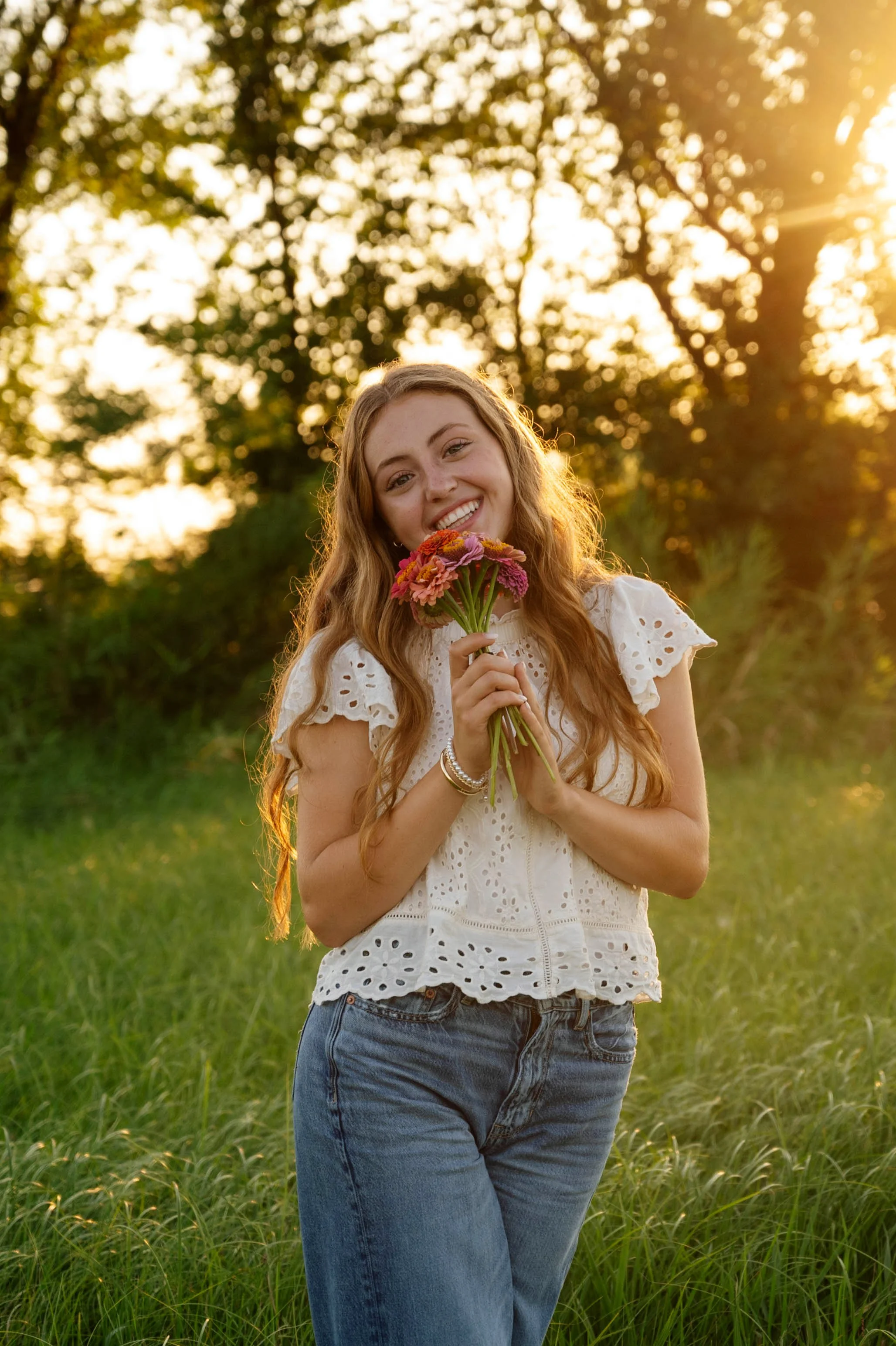 A young woman standing outdoors in a grassy area, holding a bouquet of pink and orange flowers, smiling at the camera, with sunlight filtering through trees in the background.