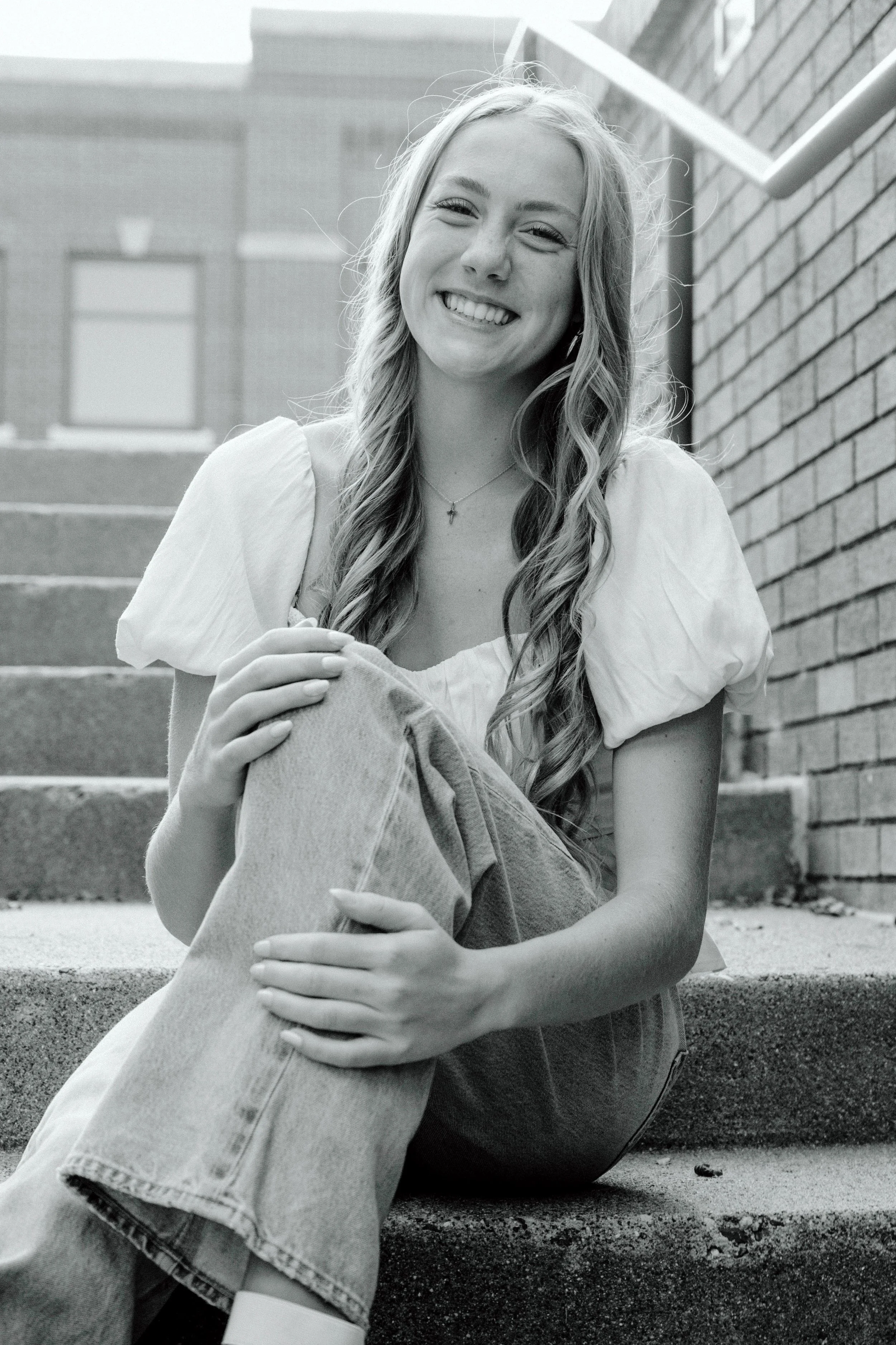 A young woman with long wavy hair, smiling and sitting on outdoor concrete stairs next to a brick wall.