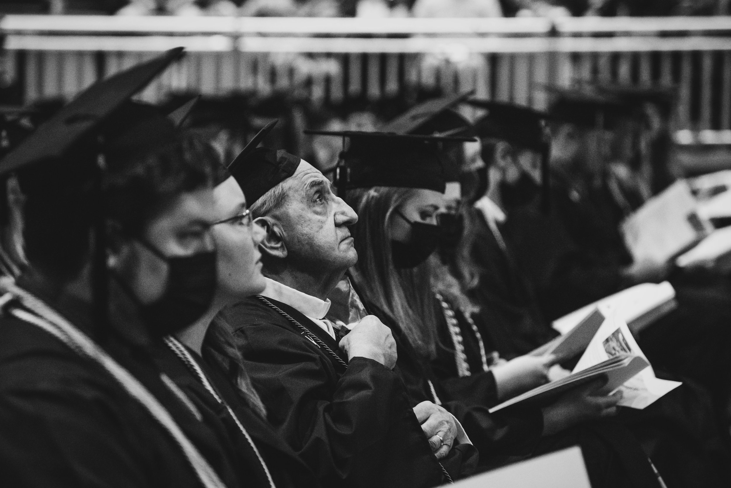 Black and white photo of graduating students seated in caps and gowns, many wearing face masks, attending a graduation ceremony.