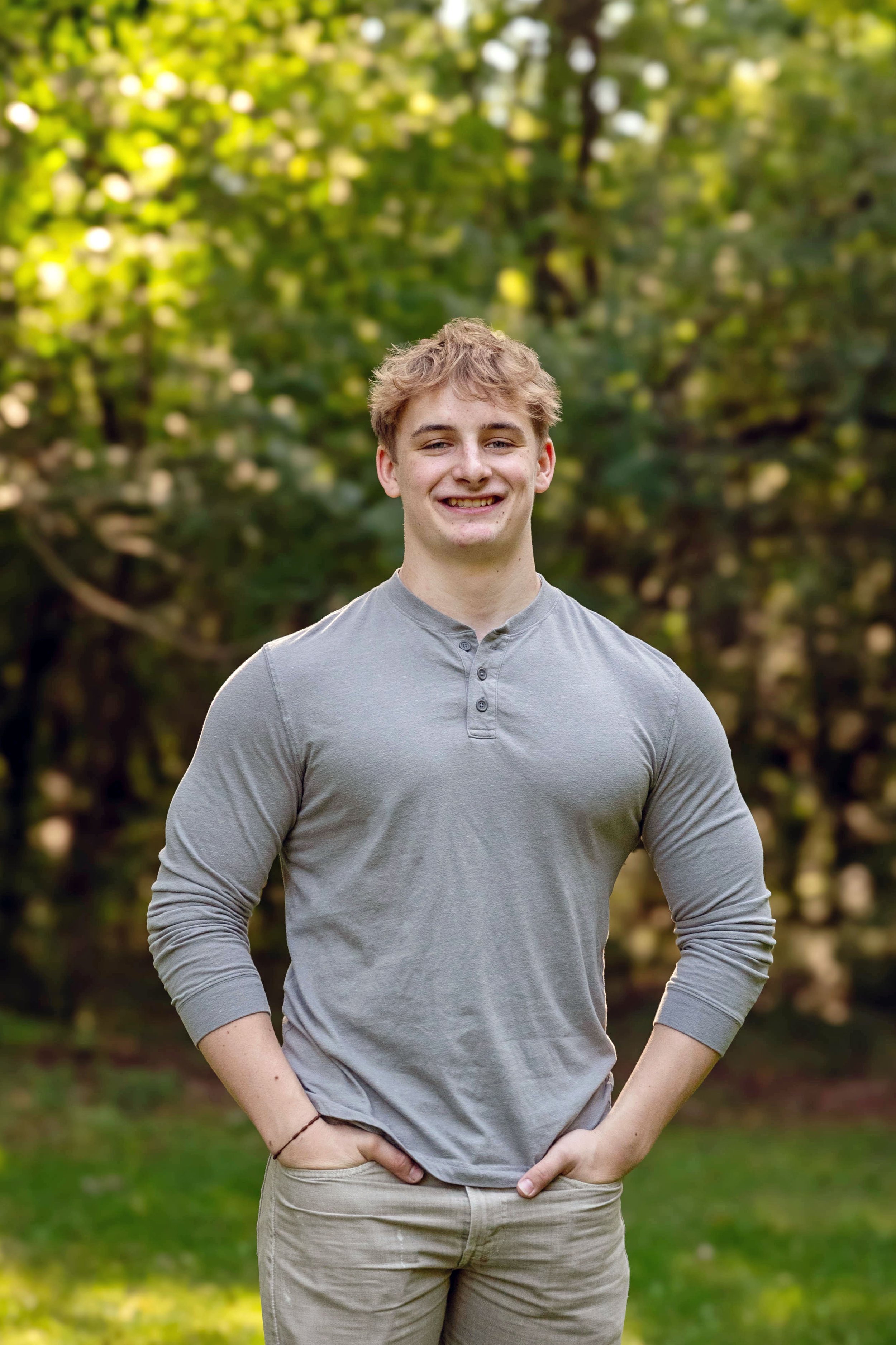 A young man standing outdoors in front of green trees, smiling, wearing a light gray long-sleeved shirt and khaki pants with his hands in his pockets.