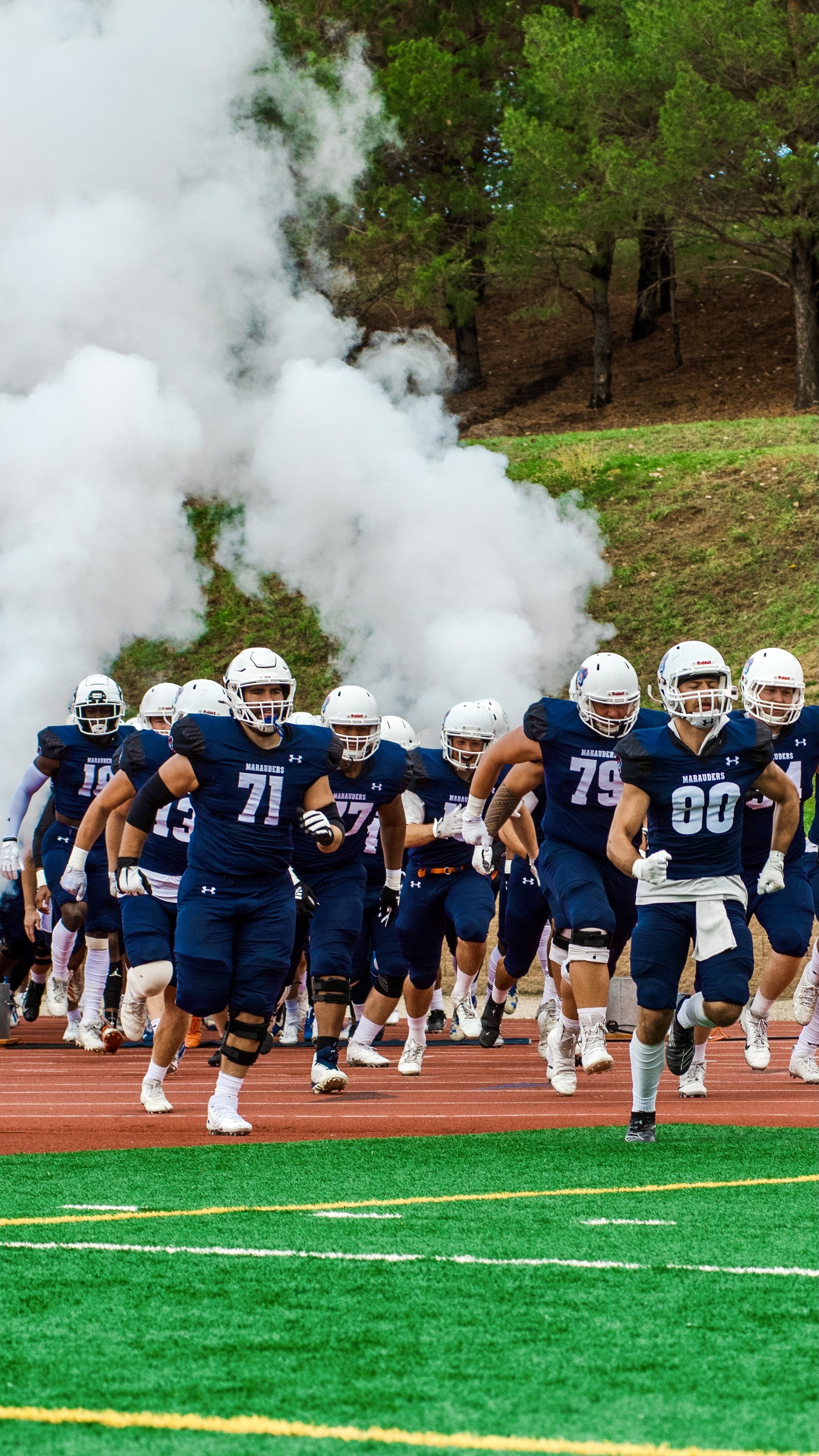 American football team in blue jerseys and white helmets running onto the field through smoke, with trees in the background.