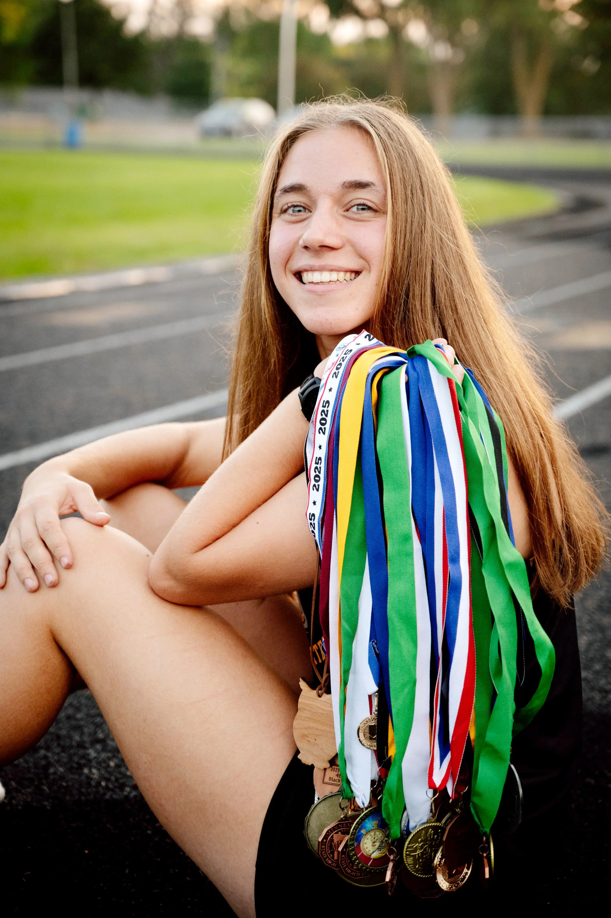 A young female athlete with long red hair sitting on a track, smiling, with multiple medals hanging around her neck, and a ribbon with '2025' printed on it.
