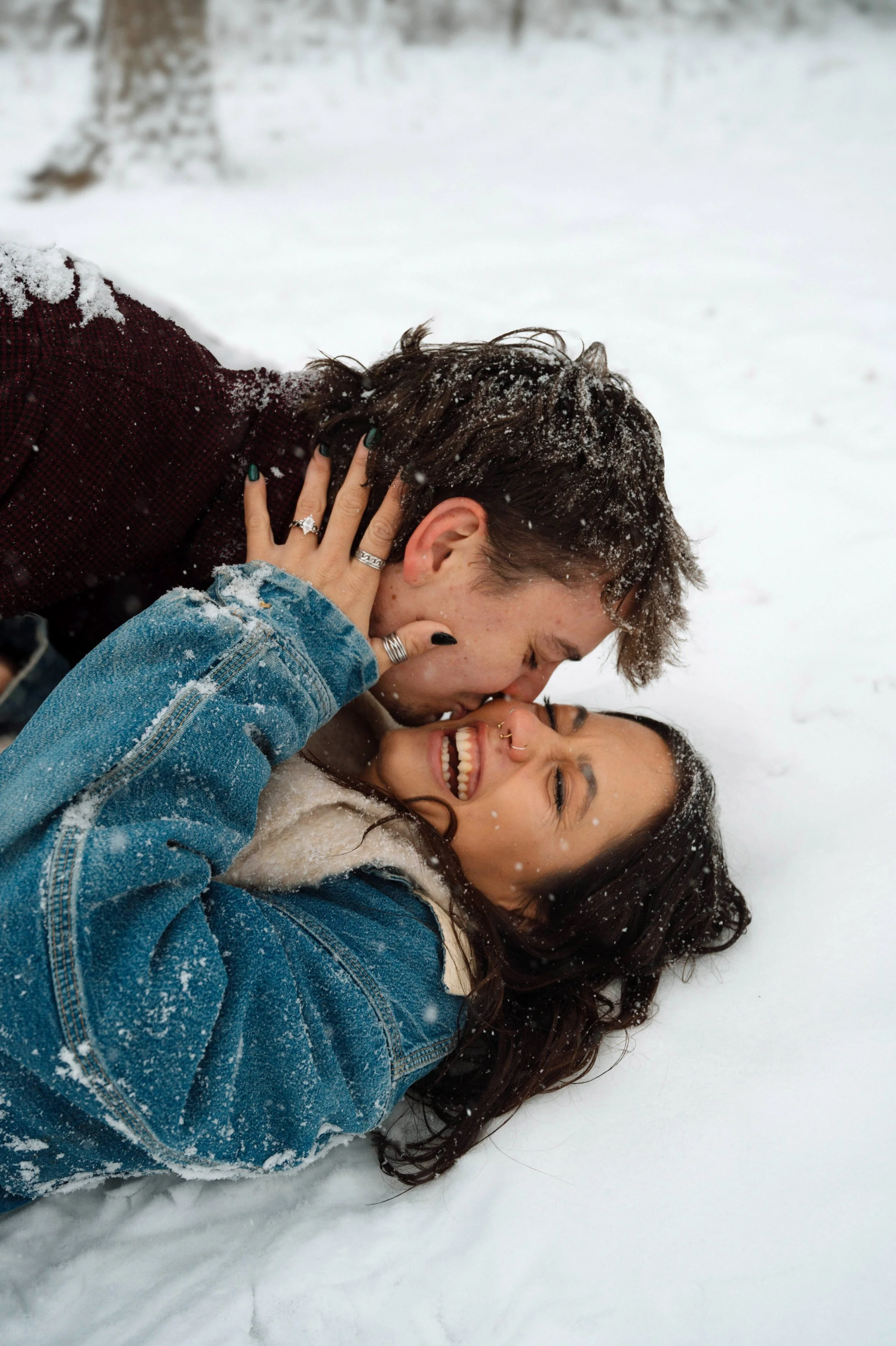 A couple lying in the snow, smiling and embracing each other, with snow on their hair and clothes, in a playful and affectionate moment outdoors.