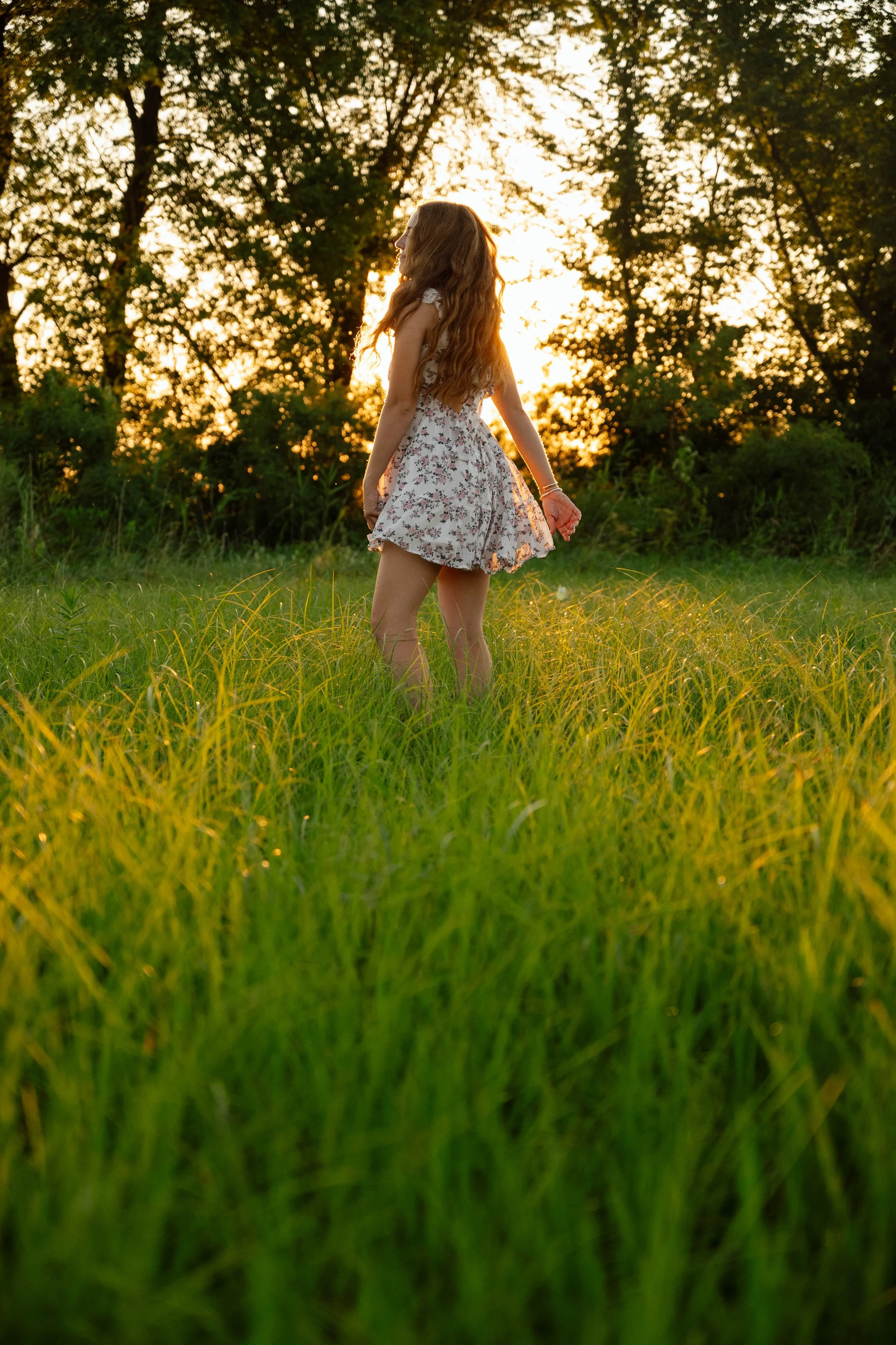 A young woman with long, wavy hair stands in a grassy field at sunset, wearing a floral dress.