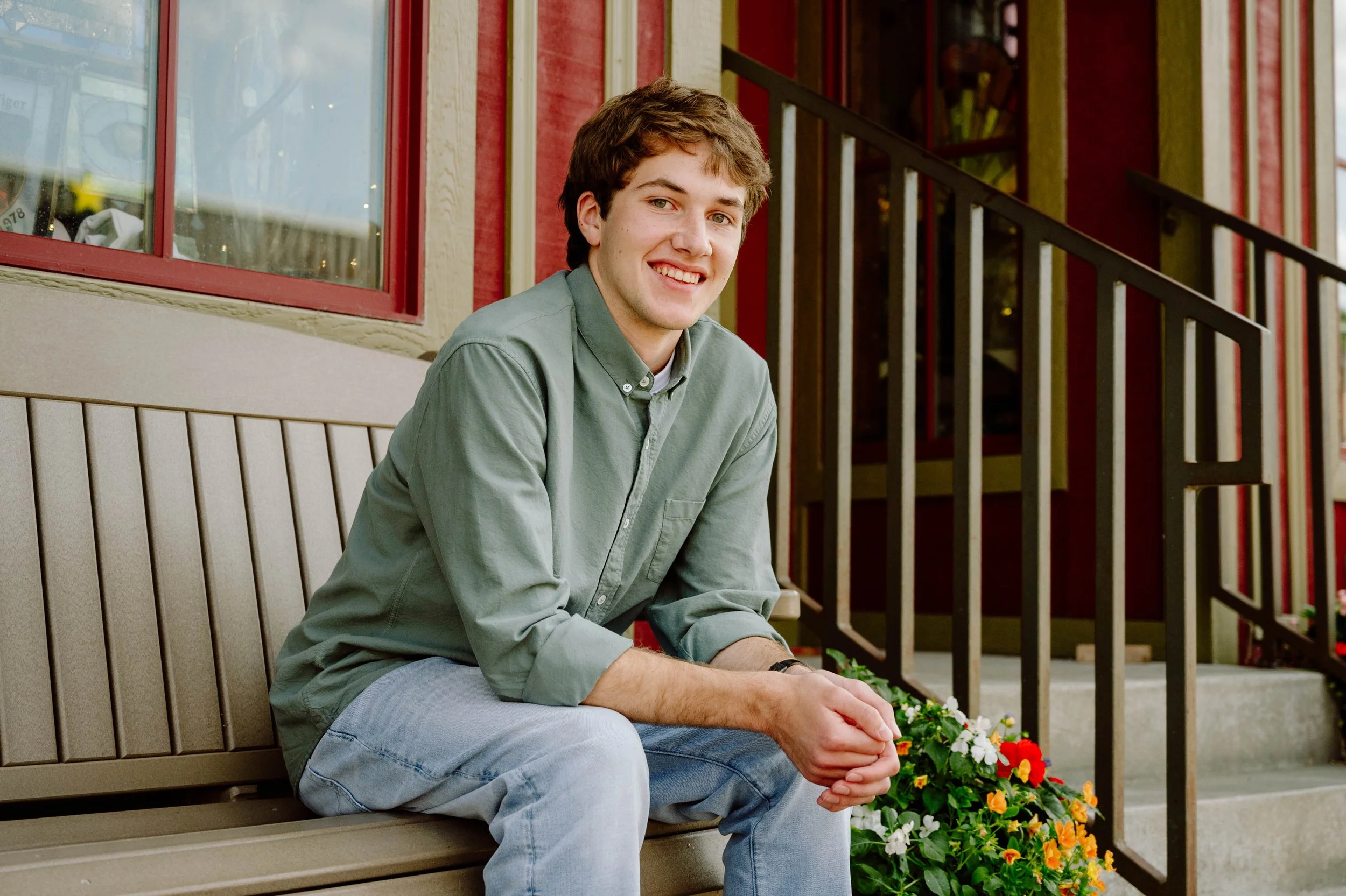 A young man with brown hair, wearing a green button-up shirt and light blue jeans, sitting on a beige bench outside a building with red window frames and a flower bed with white, orange, and yellow flowers, smiling at the camera.