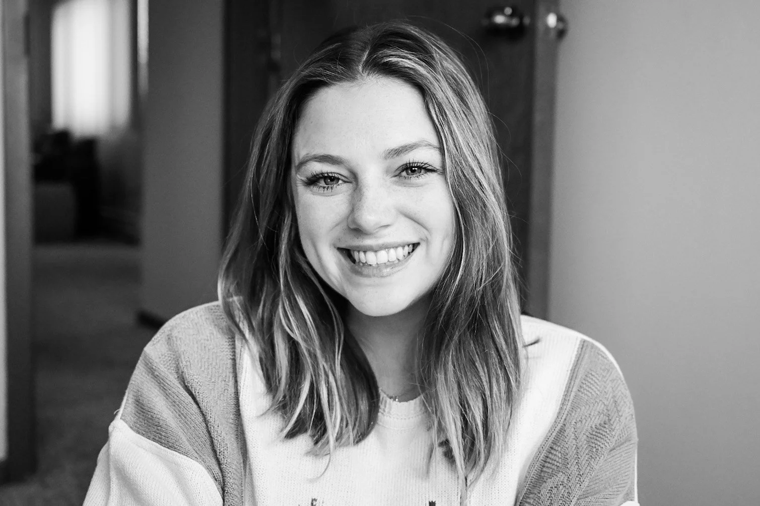 A young woman smiling at the camera, in a black and white photo. She has shoulder-length hair and is wearing a casual sweater. The background shows an indoor setting with furniture.