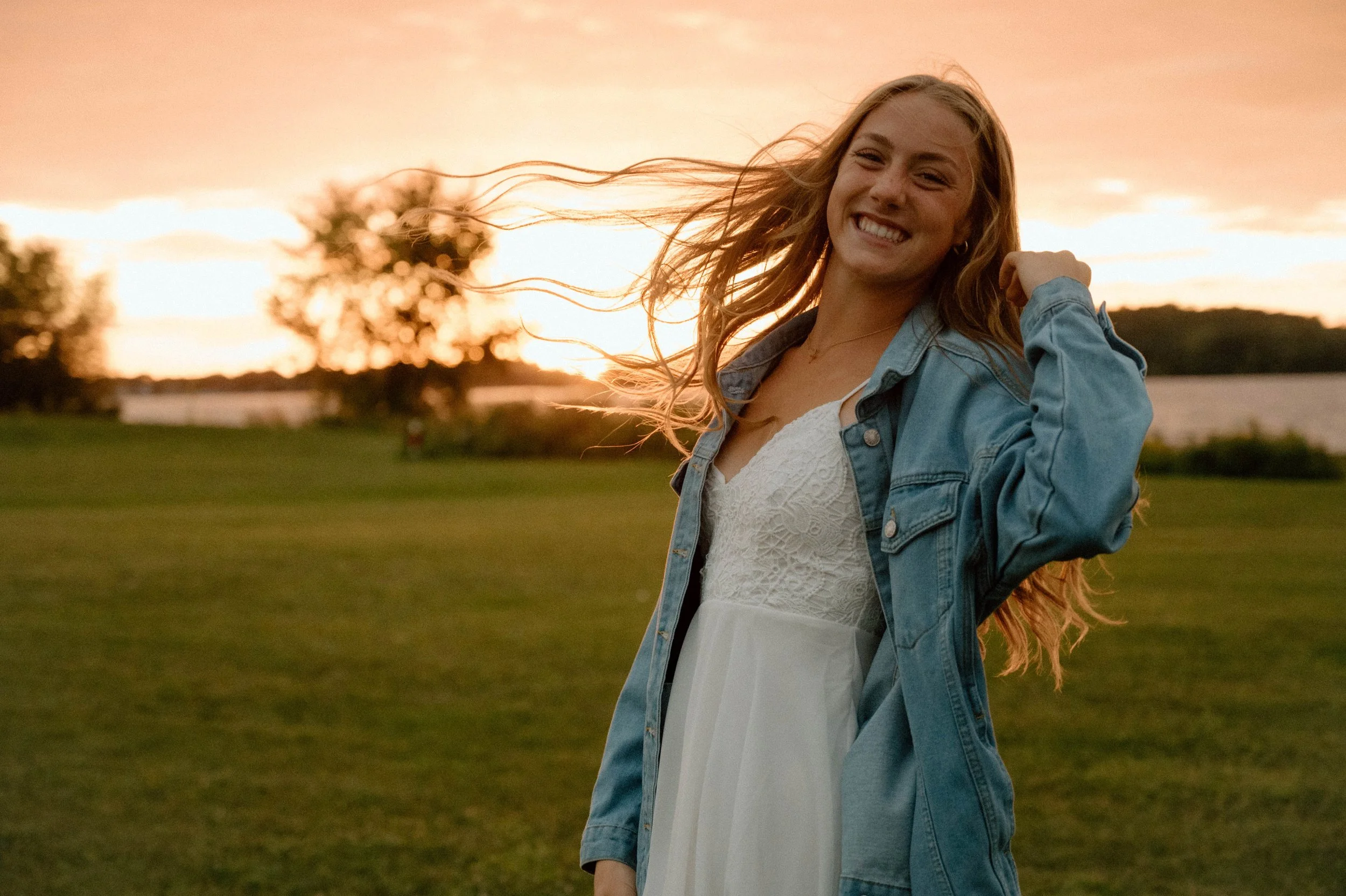 A young woman with long blonde hair smiling and posing outdoors during sunset, wearing a white dress and a denim jacket, with wind blowing her hair.