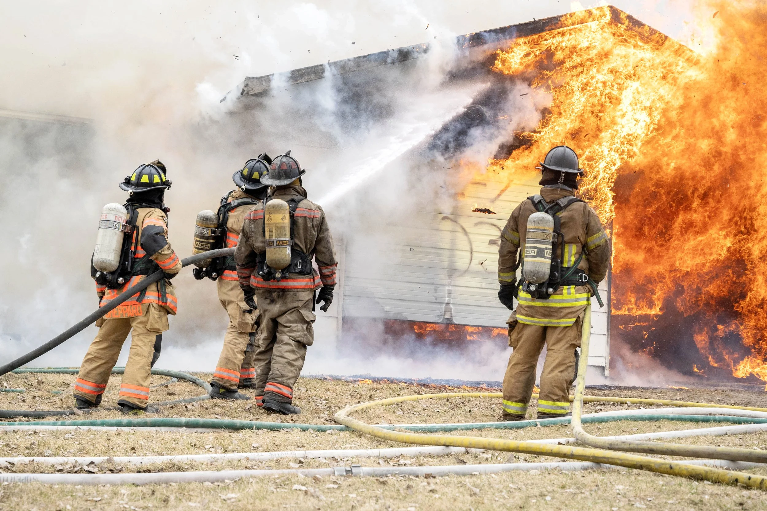 Four firefighters in firefighting gear working to extinguish a house fire with flames and smoke engulfing the building.