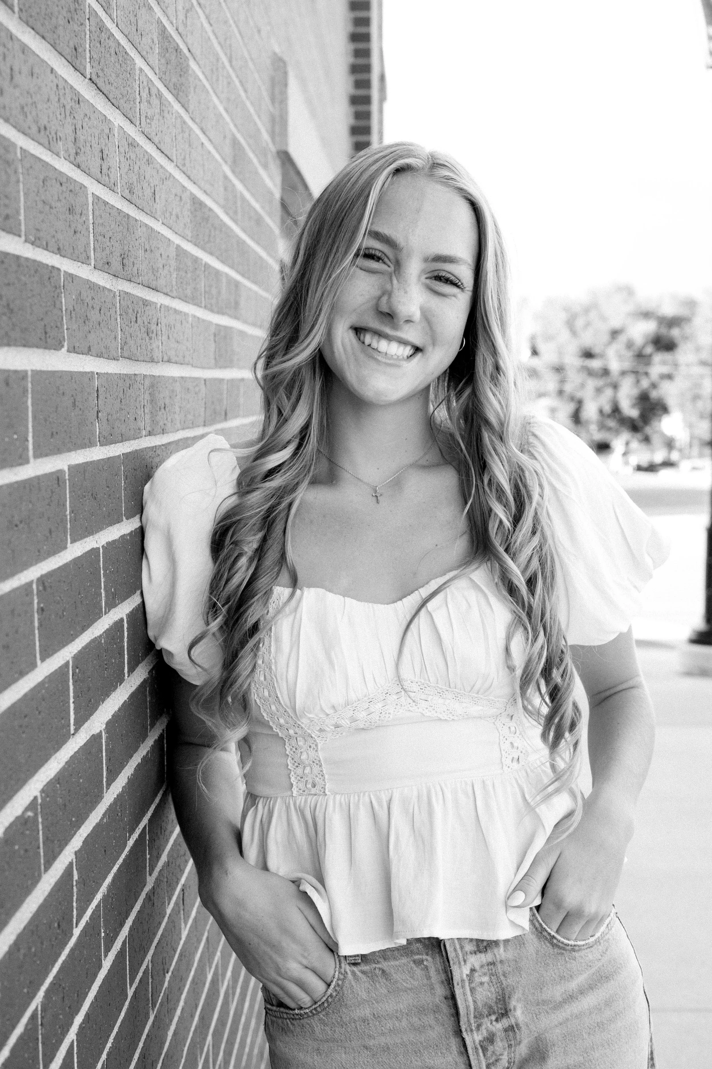 Black and white photo of a smiling young woman with long wavy hair, standing outdoors leaning against a brick wall, wearing a short-sleeved, ruffled blouse and jeans.