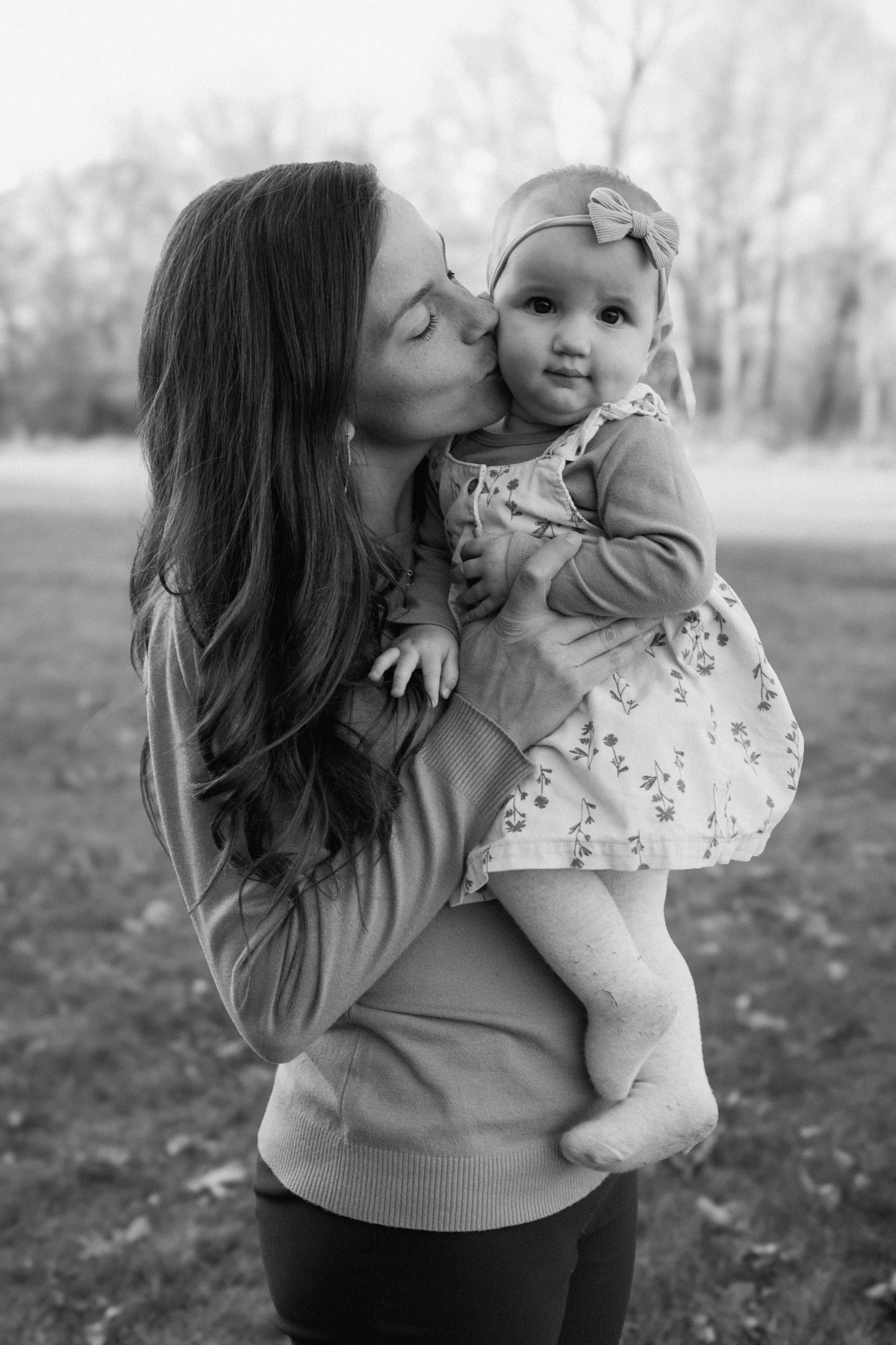 A woman holding a young girl outdoors, with the woman kissing the girl on the cheek in a black-and-white photo.