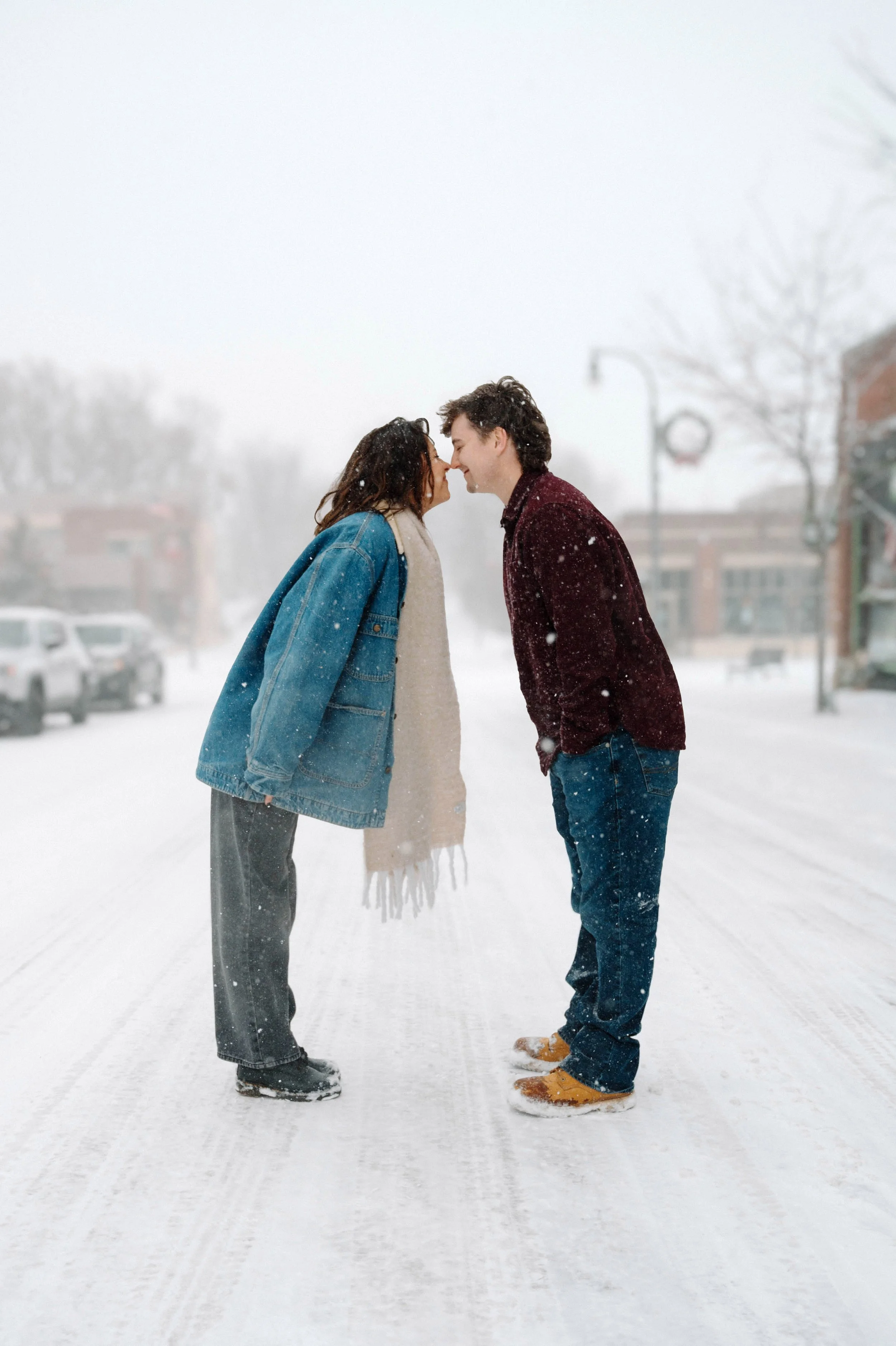 A couple standing close in a snowy street, touching foreheads and smiling, with snow falling around them.