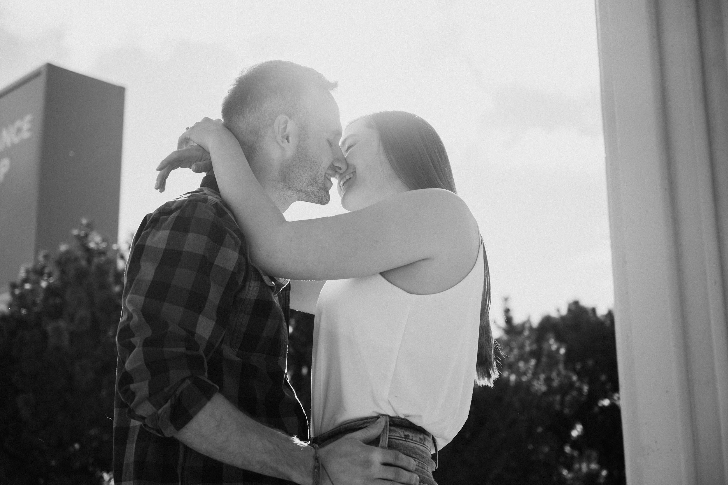 A black and white photo of a couple standing close, touching foreheads and smiling with their eyes closed, outdoors in natural light.