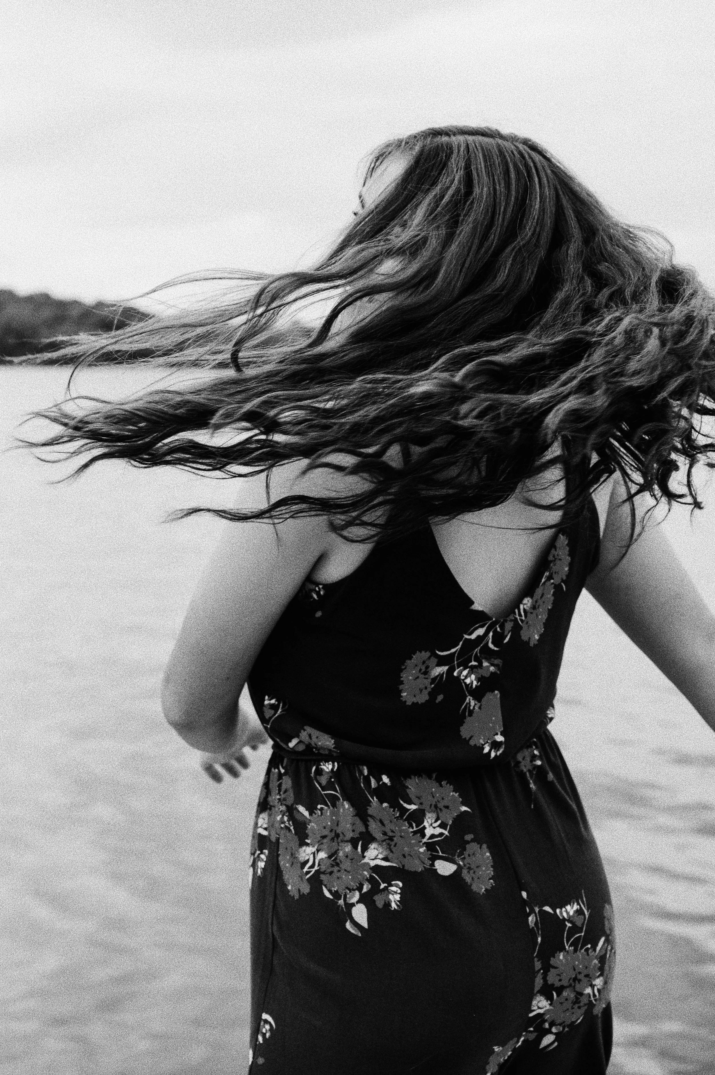 A woman with long, wavy hair flowing in the wind, wearing a sleeveless floral dress, near water.