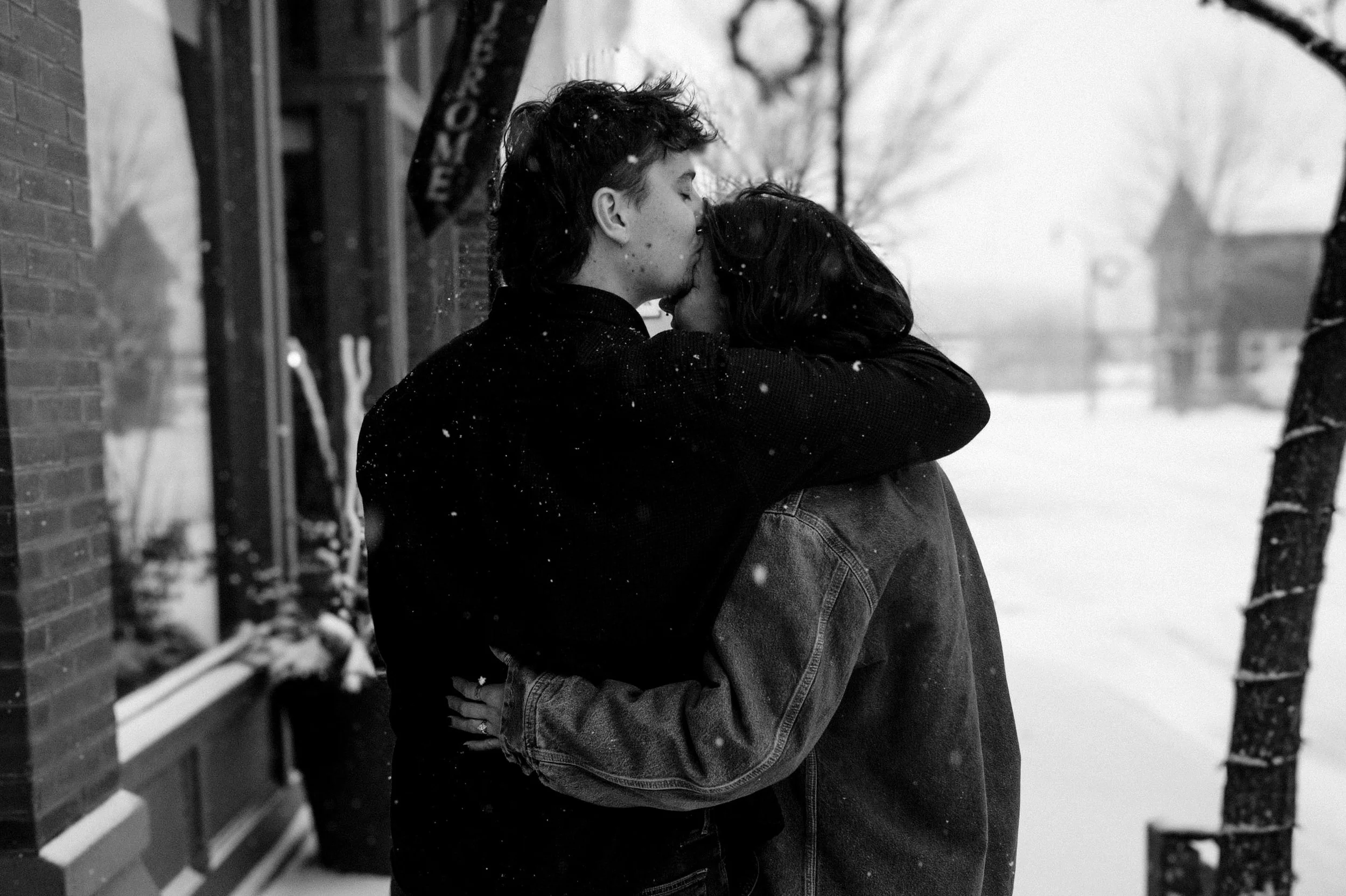 A black and white photo of a couple hugging each other outside in a snowy setting. The man is kissing the woman's forehead, and snow is falling.