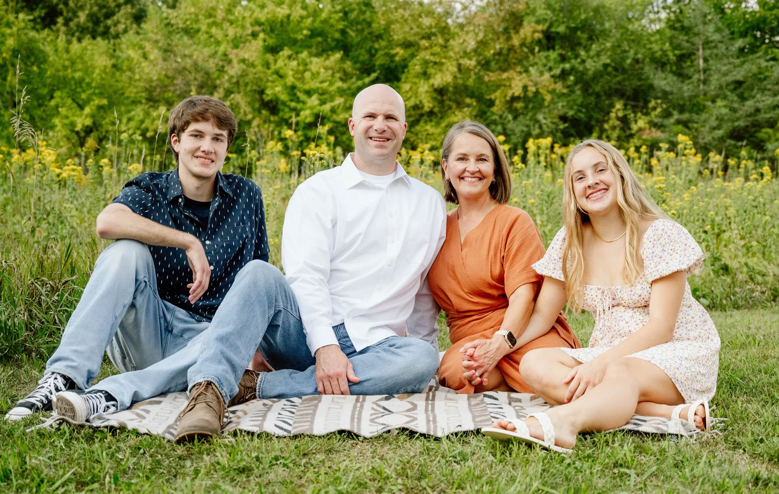 A family of four sitting on a blanket outdoors in a grassy field with yellow flowers and green trees in the background.