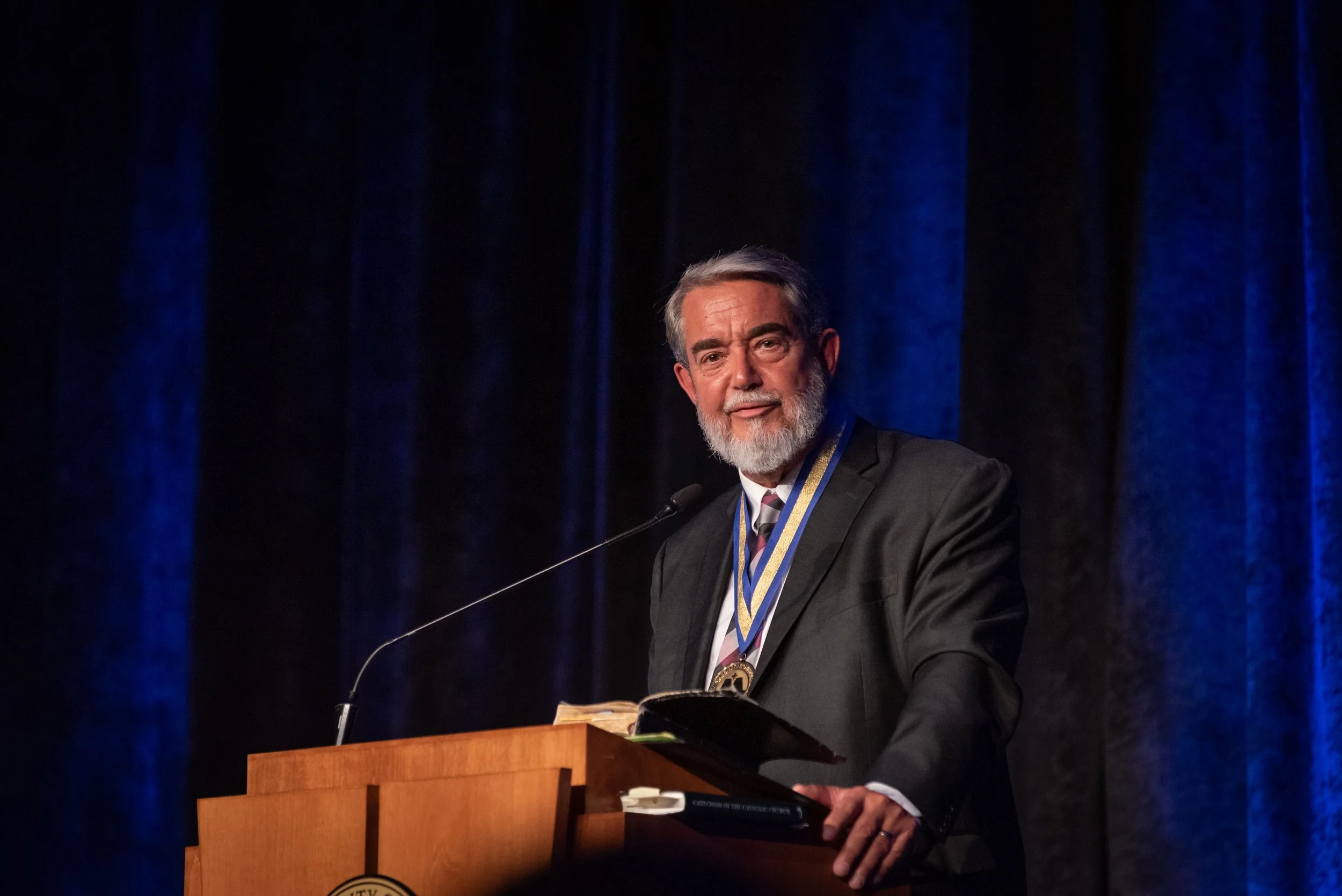 An elderly man with gray hair and beard, wearing a dark suit and tie, stands at a wooden podium with a microphone, wearing a medal around his neck, in front of blue stage curtains.