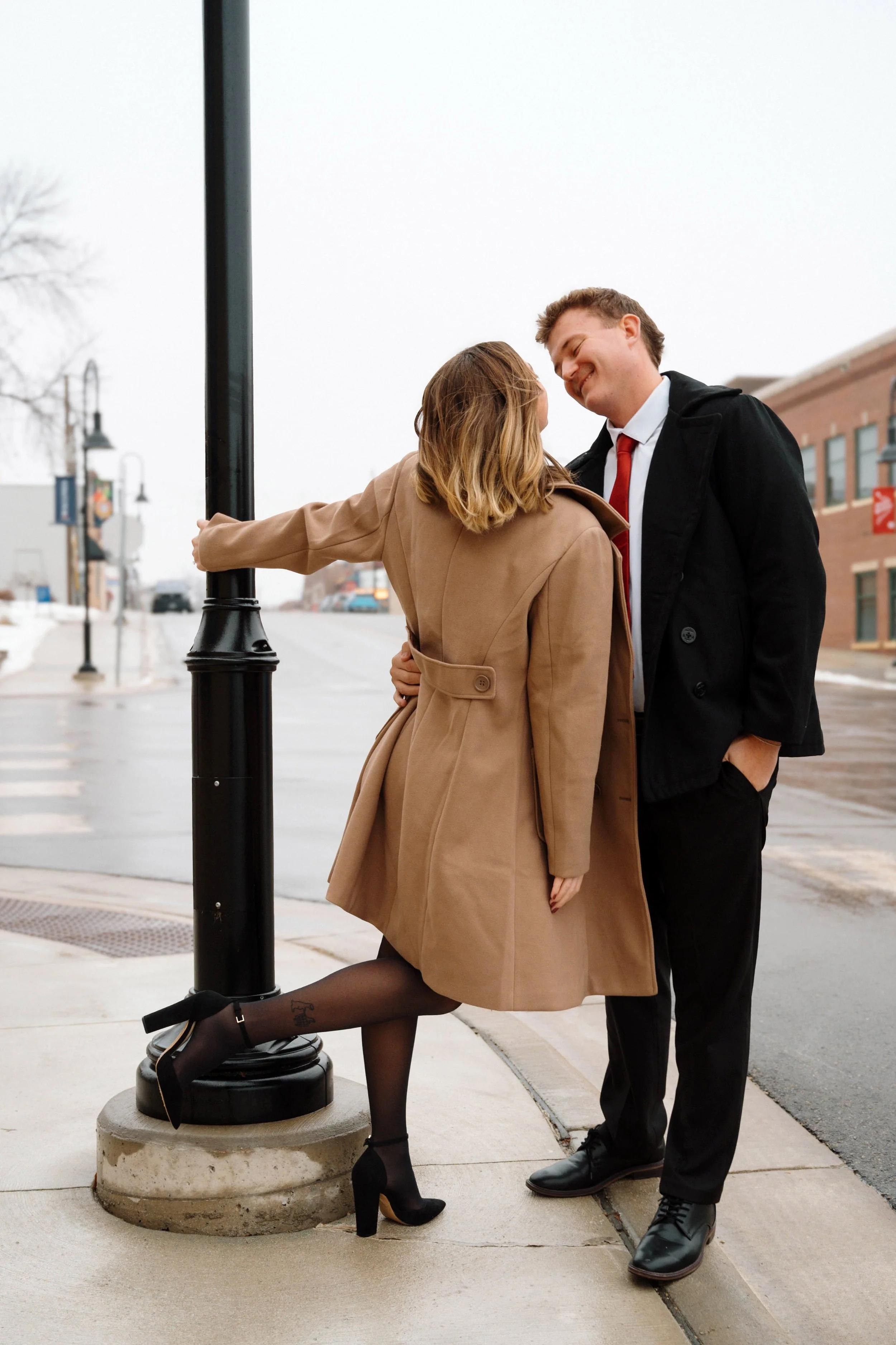 A man and woman standing on a city sidewalk, smiling and leaning close to each other, with the woman's arm resting on a streetlamp.