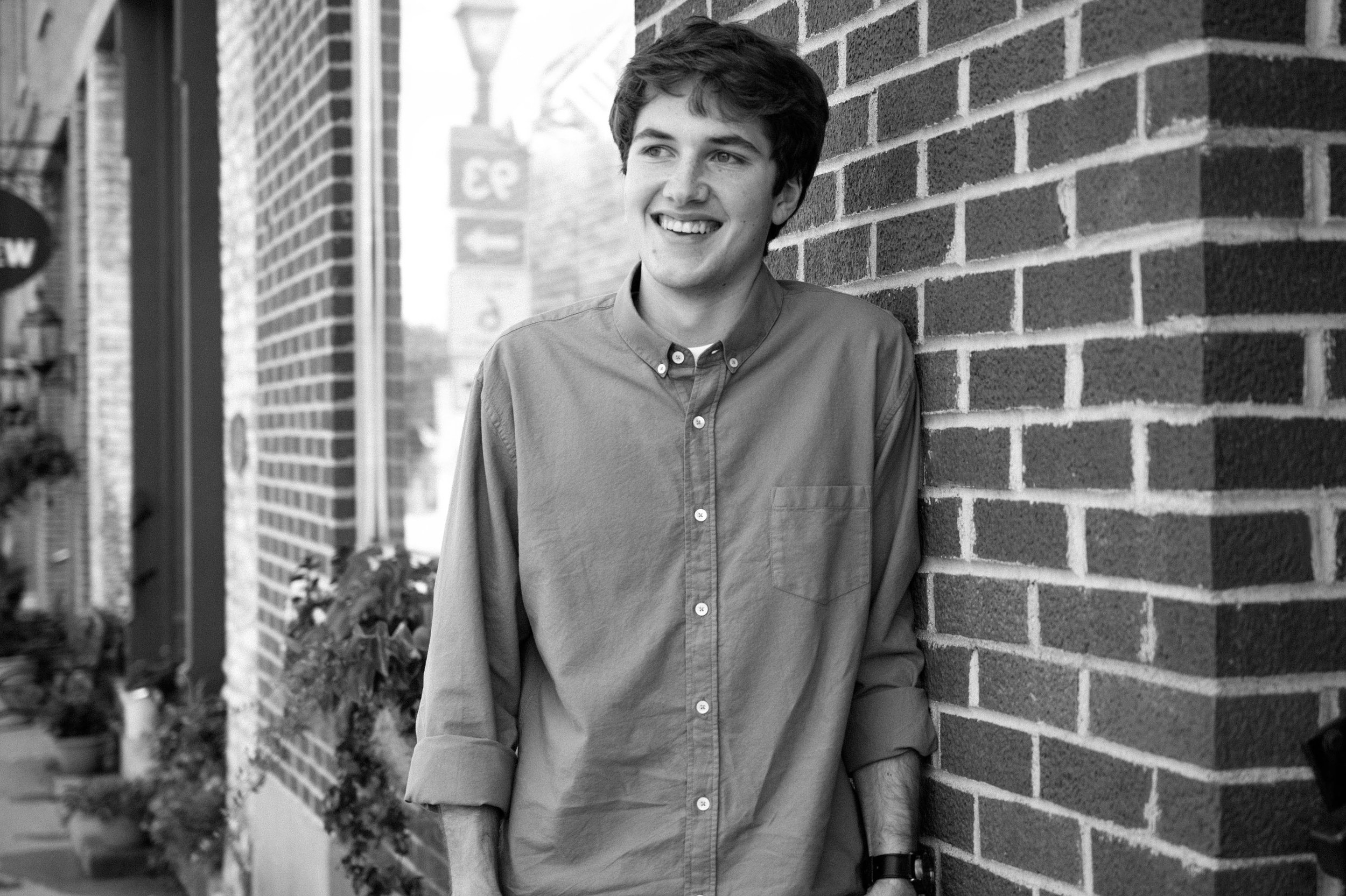 A young man leaning against a brick wall, smiling, with shops and potted plants visible in the background.