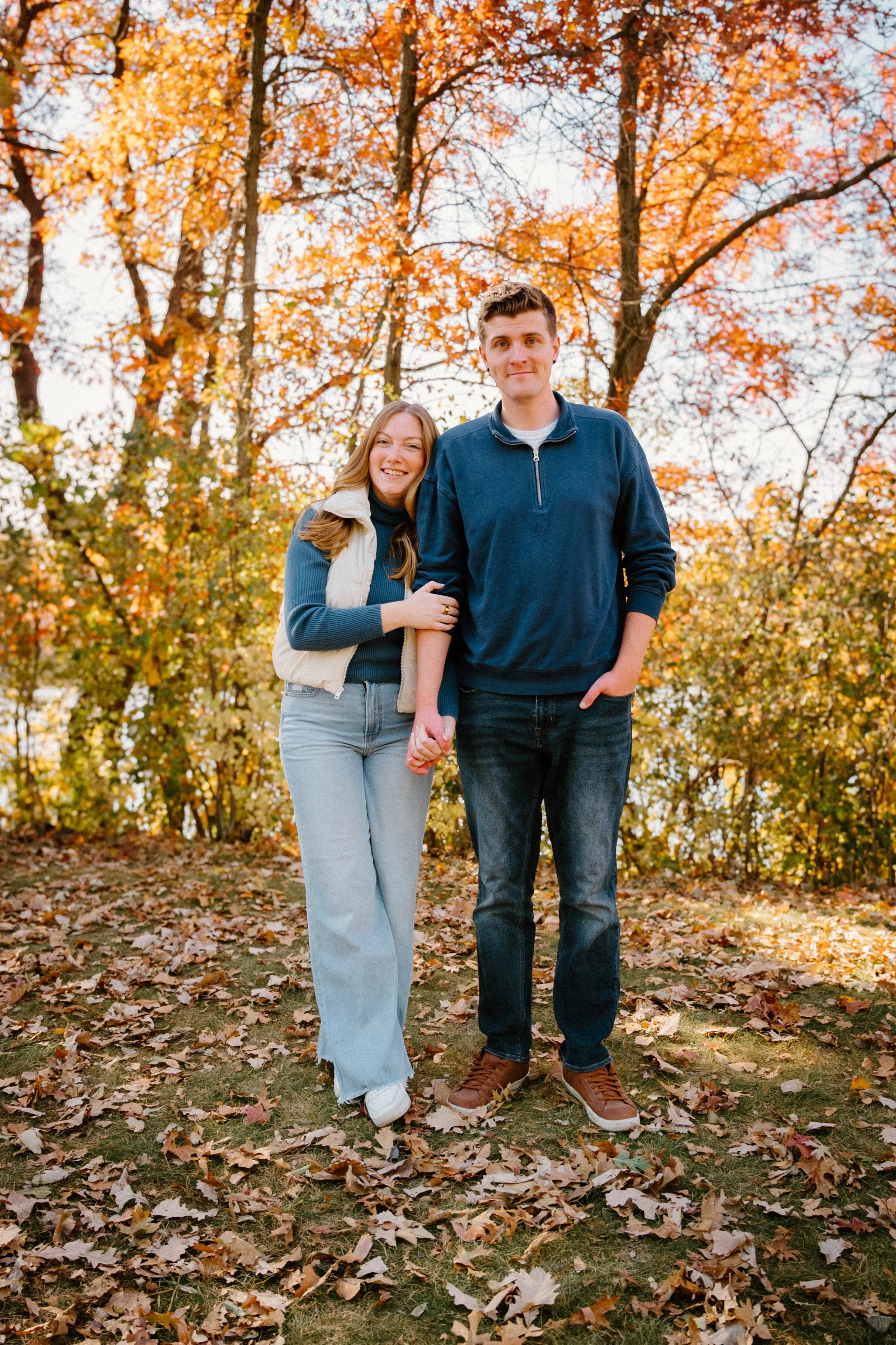 A young couple walking hand-in-hand outdoors during fall, surrounded by colorful autumn leaves and trees.