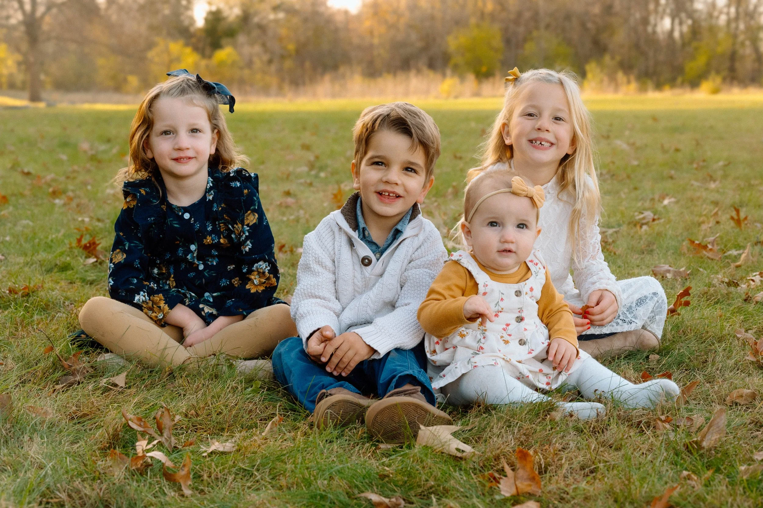 Four children, two girls and two boys of different ages, sitting on the grass in an outdoor park during autumn, with fallen leaves and trees in the background.
