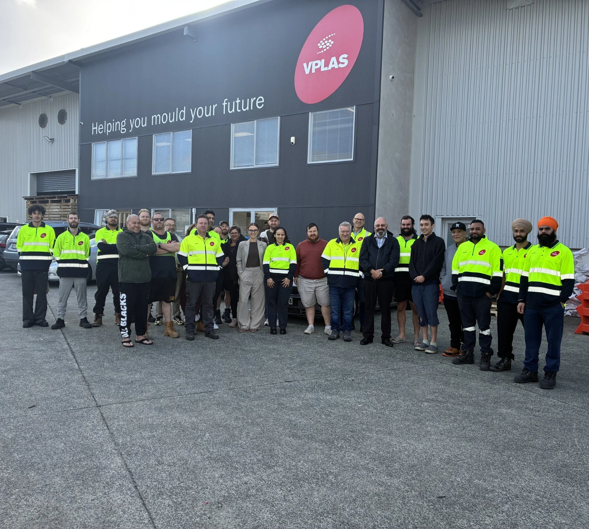 Group of people standing in front of a building with a sign that reads 'Helping you mould your future' and the logo 'VPLAS', some wearing high-visibility training vests.