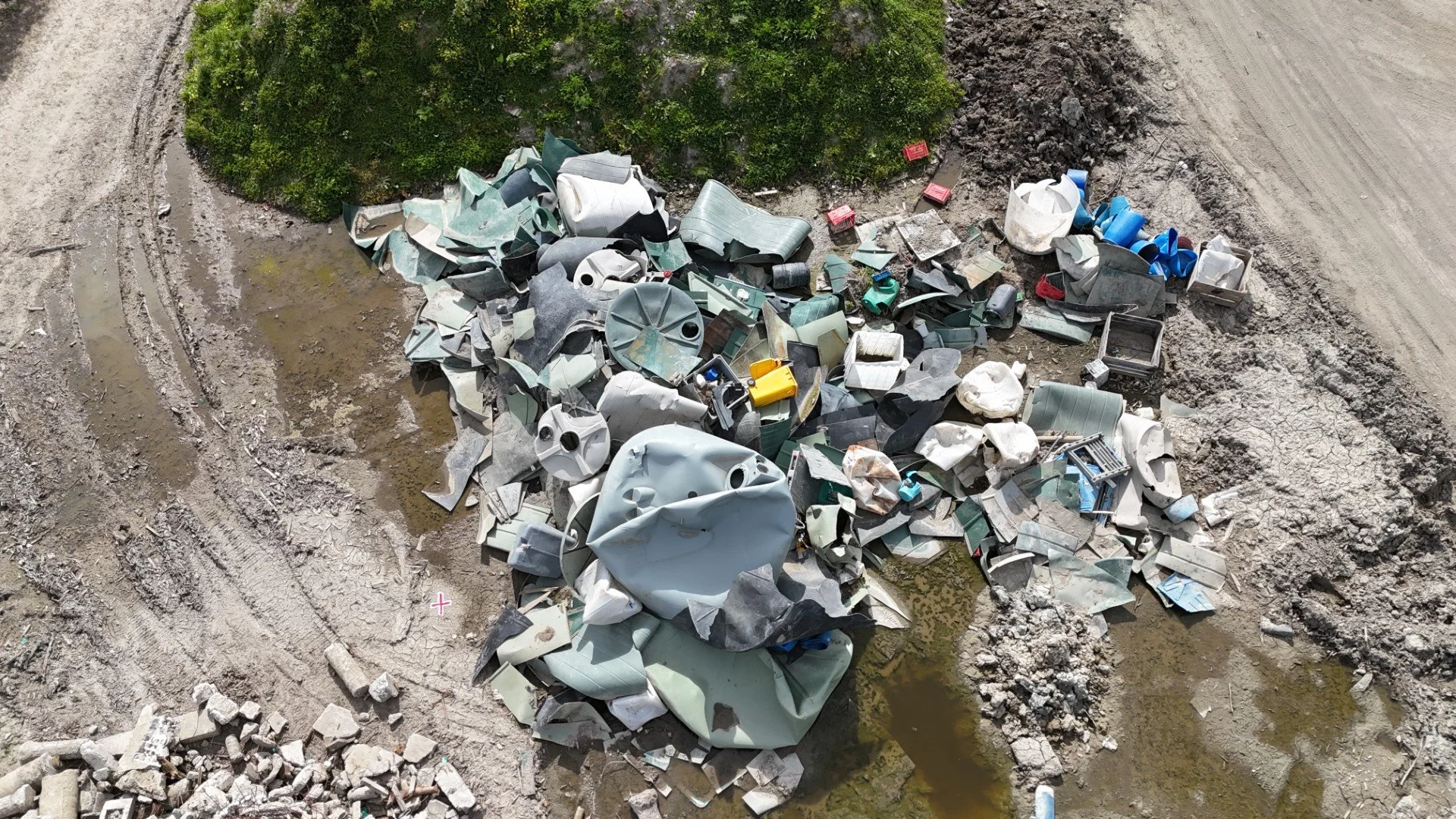 Aerial view of a landfill with various discarded construction materials, debris, and garbage piled on muddy ground near a patch of green vegetation.