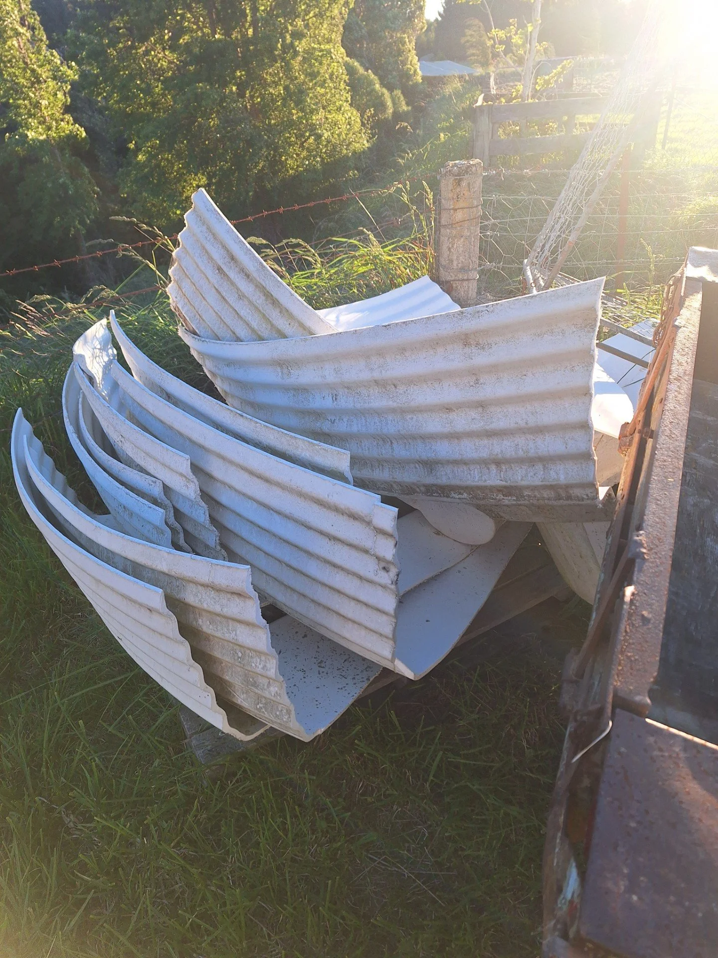 A pile of white, corrugated metal roofing sheets leaning against a weathered wooden fence outdoors, with green trees and sunlight in the background.