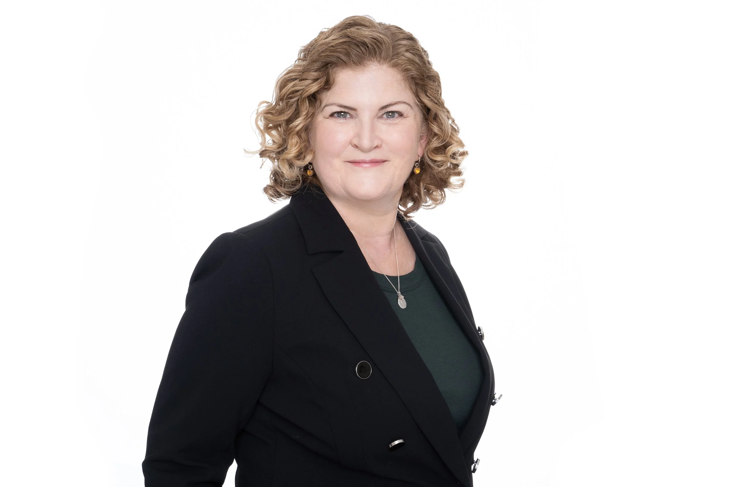 A middle-aged woman with shoulder-length curly blonde hair, wearing a black blazer over a dark green top, gold earrings, and a silver necklace, standing against a white background and looking at the camera with a slight smile.