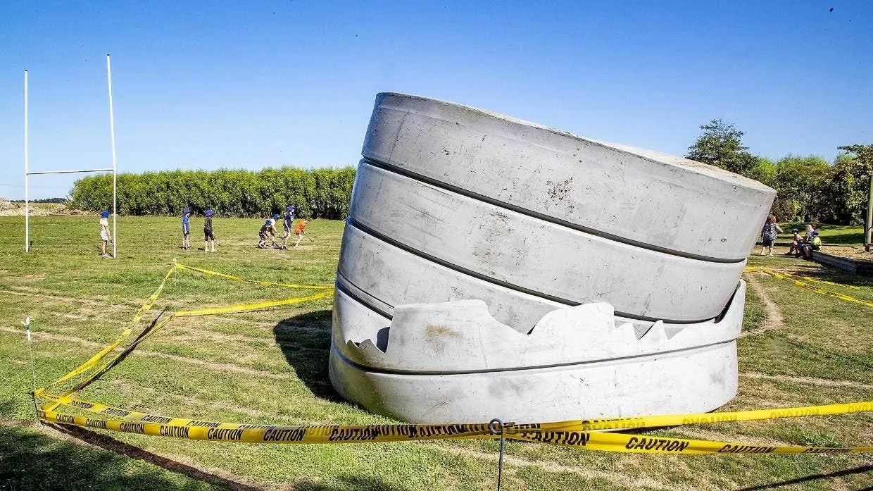 Large sculpture resembling a fractured head with closed eyes, made of concrete or stone, on a grassy field with yellow caution tape around it. In the background, children in blue caps are playing sports on a field during daytime with clear blue sky.