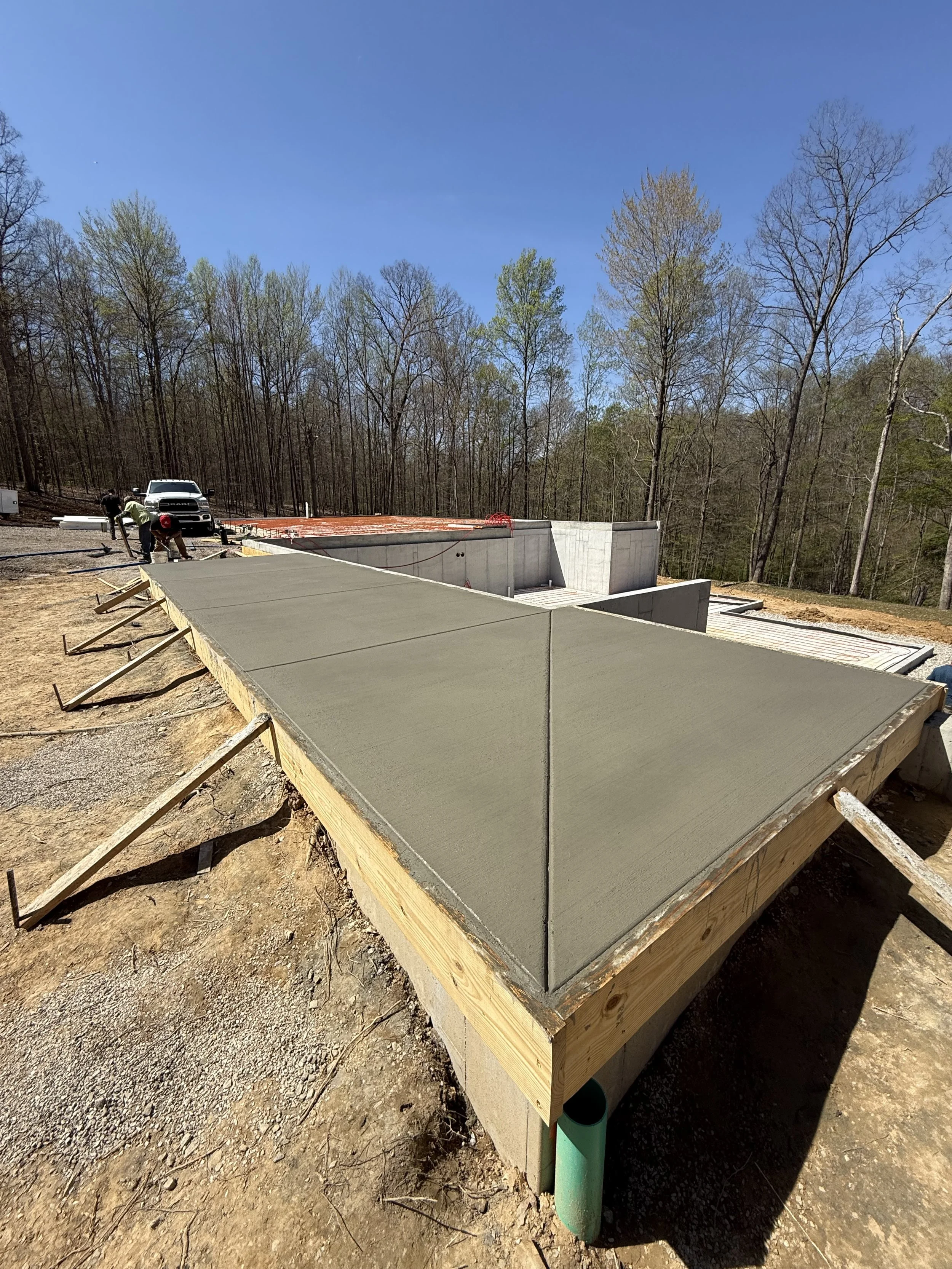 Concrete foundation being poured for a building, with construction workers and a vehicle in the background, and trees on a clear, sunny day.