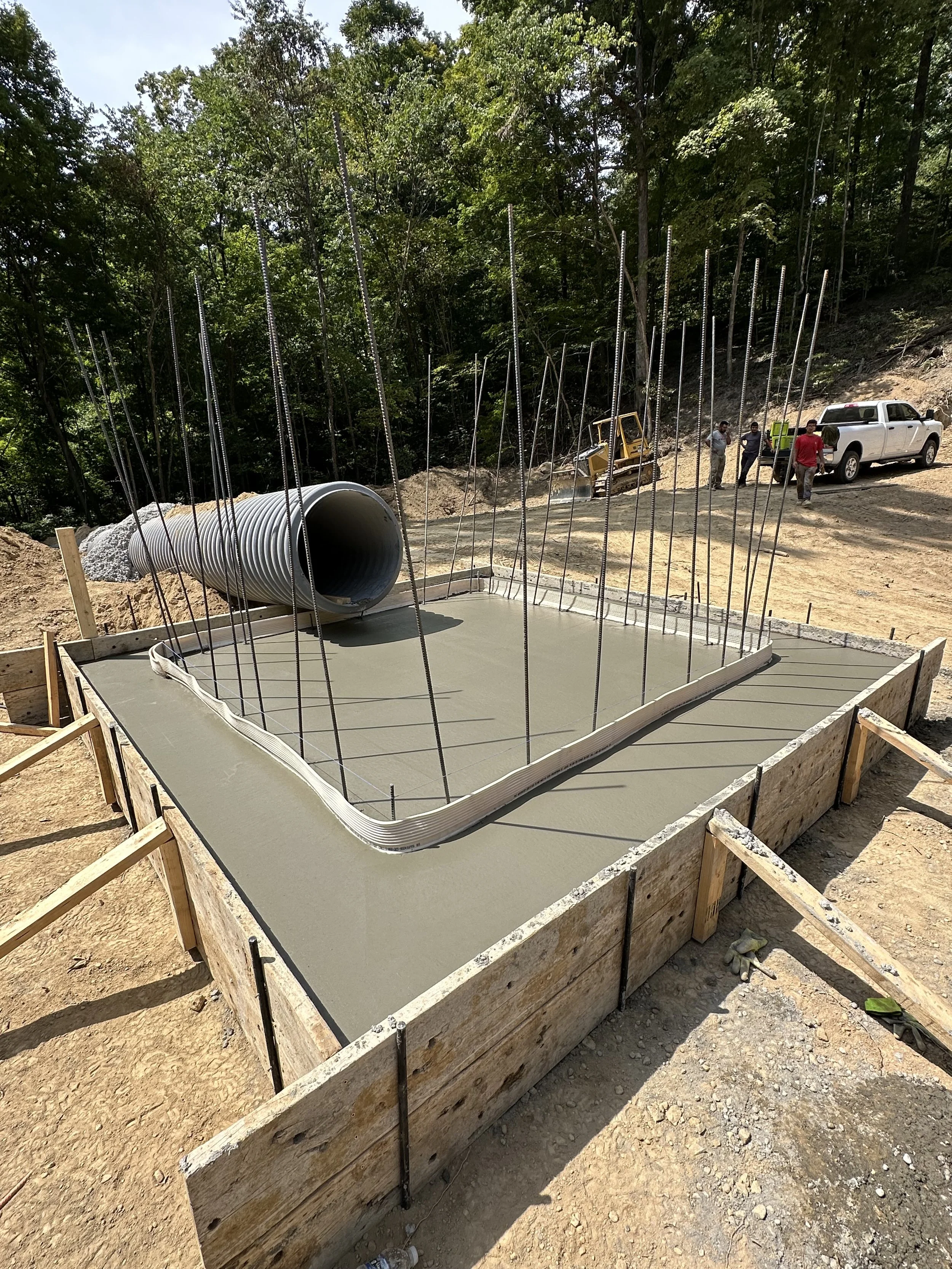 Construction site with a freshly poured concrete foundation, metal rebar protruding, a large drainage pipe, and construction workers and vehicles in the background amidst trees.