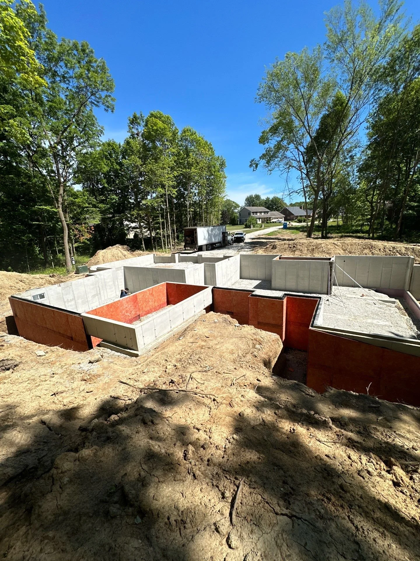 Construction site with concrete foundation walls for a house, surrounded by dirt and trees, under a bright blue sky.
