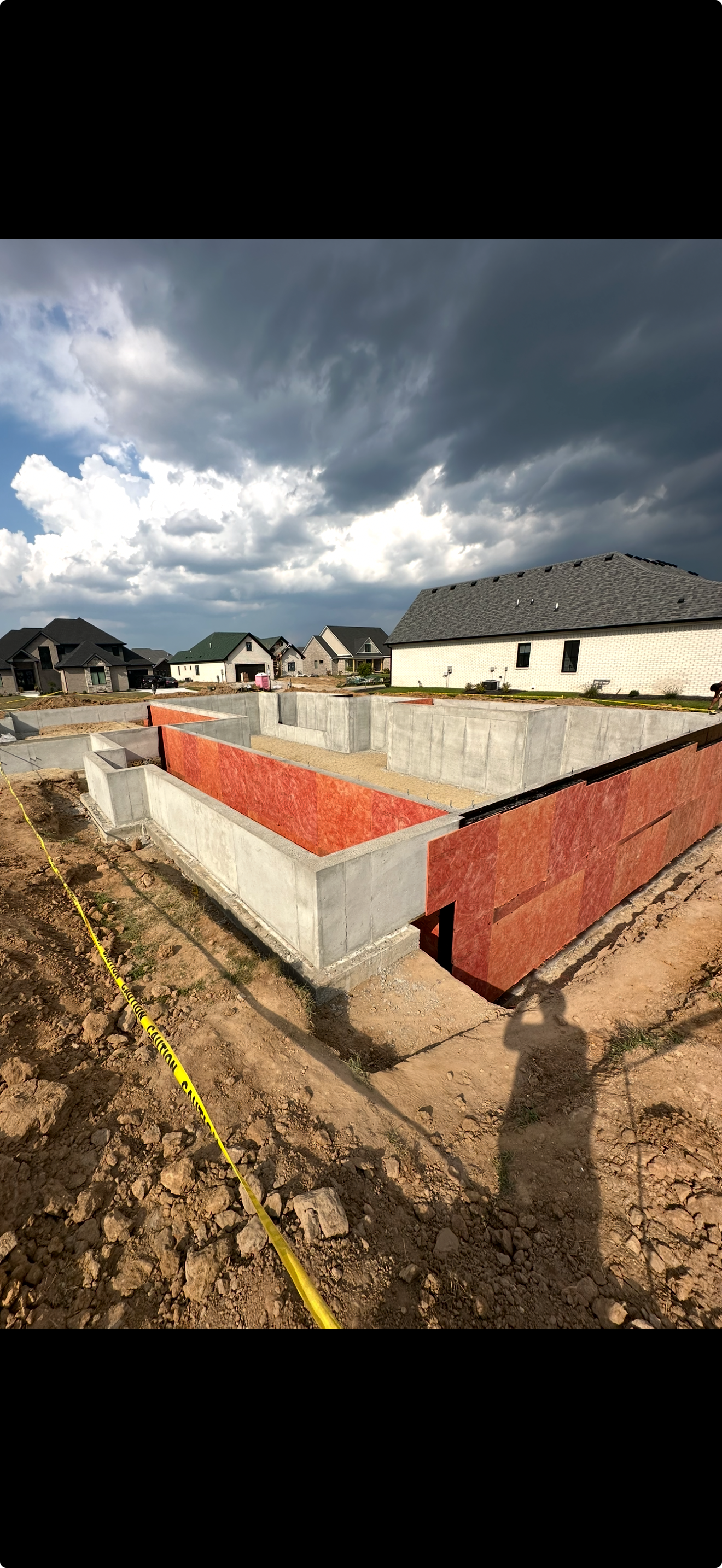 Construction site with concrete foundation walls and red sheathing, under a dark, cloudy sky.