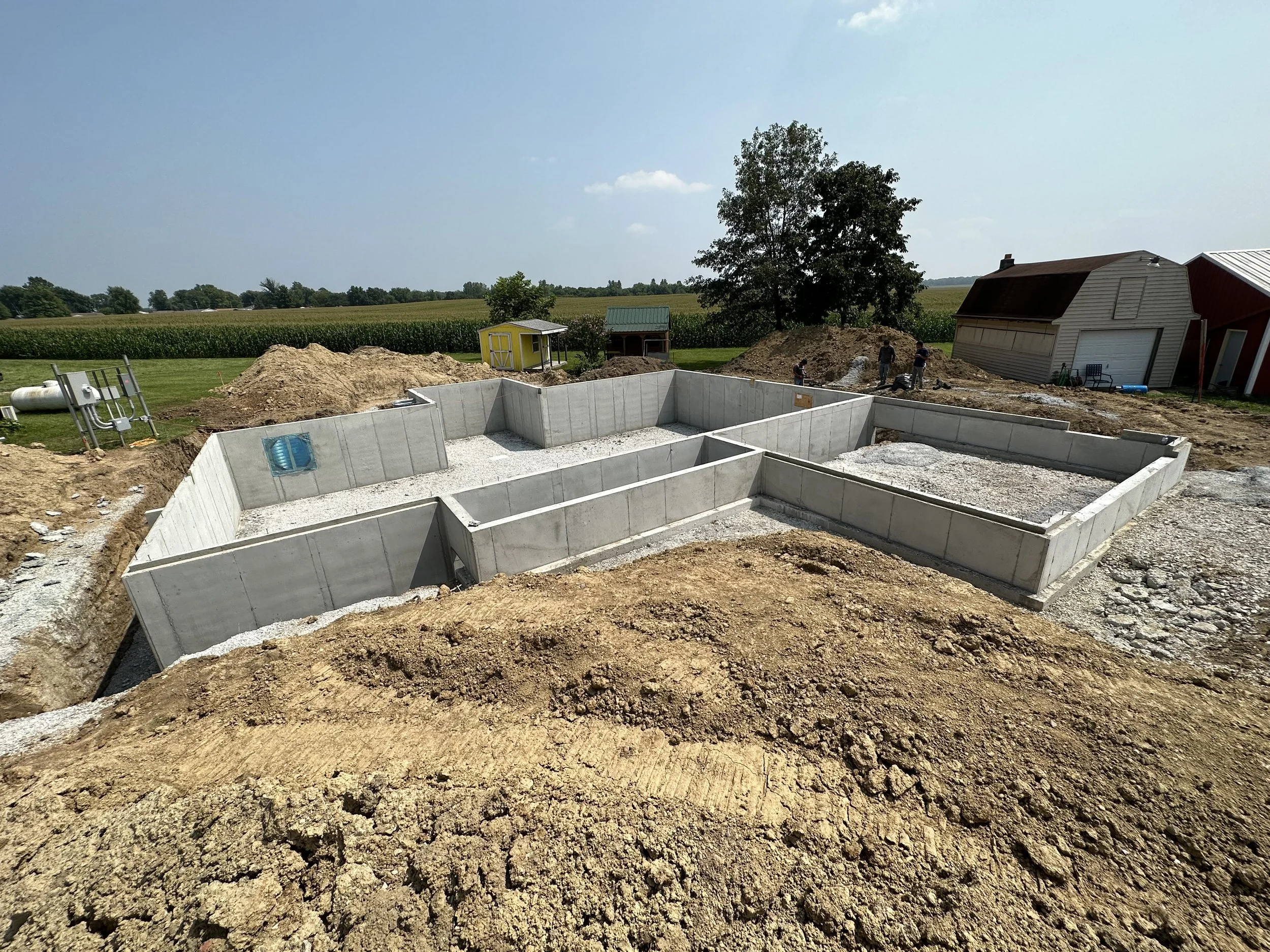 Construction site with concrete foundation walls for a building, surrounded by dirt and dirt piles, with a rural landscape in the background including a barn, trees, and fields under a clear blue sky.