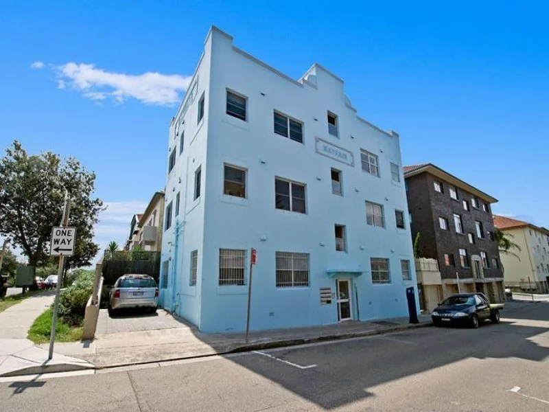 Light blue multi-story apartment building on a city street with cars parked outside and a one-way street sign nearby.