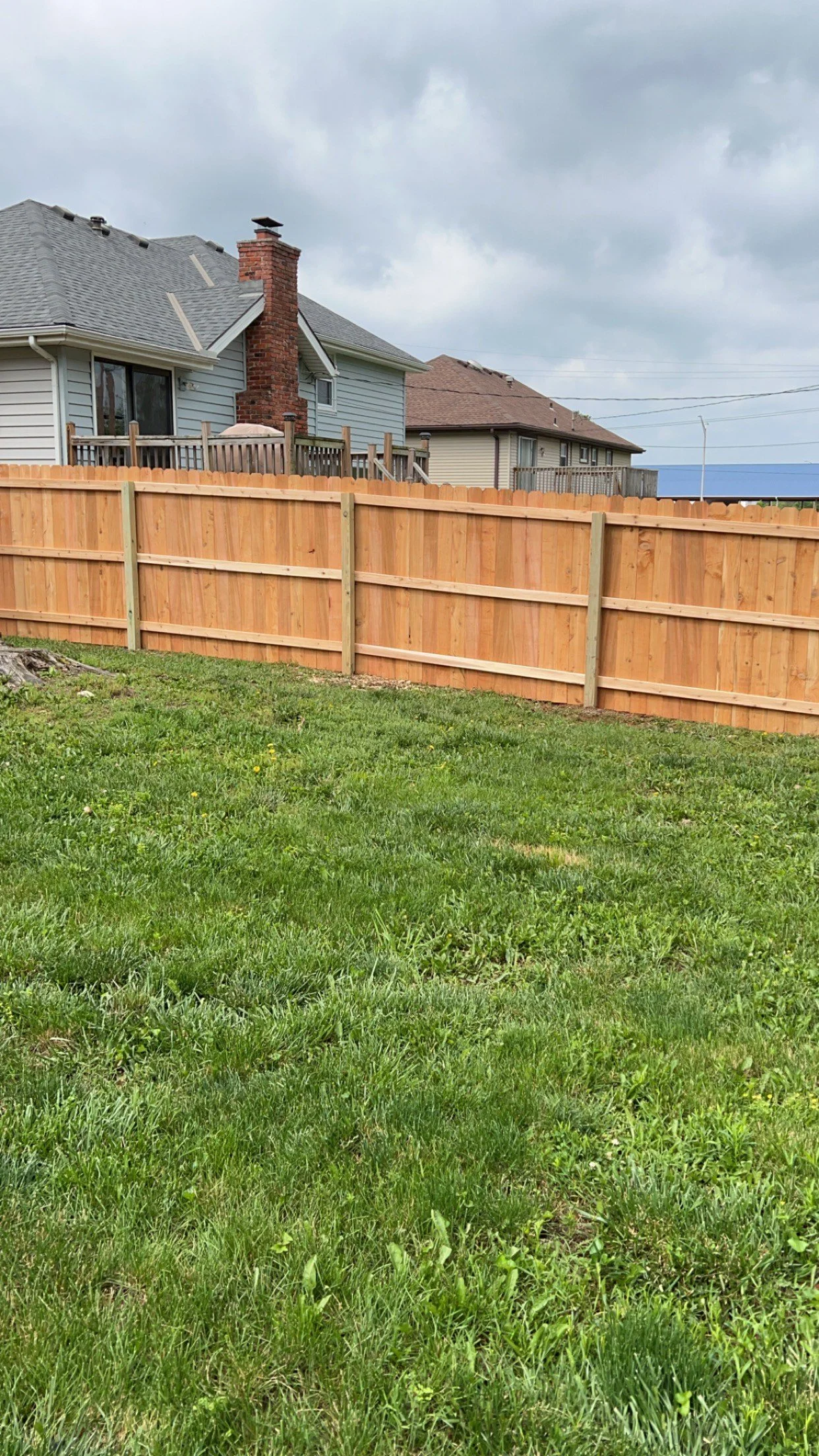 A backyard with green grass, a wooden fence, and neighboring houses with rooftops visible in the background under a cloudy sky.