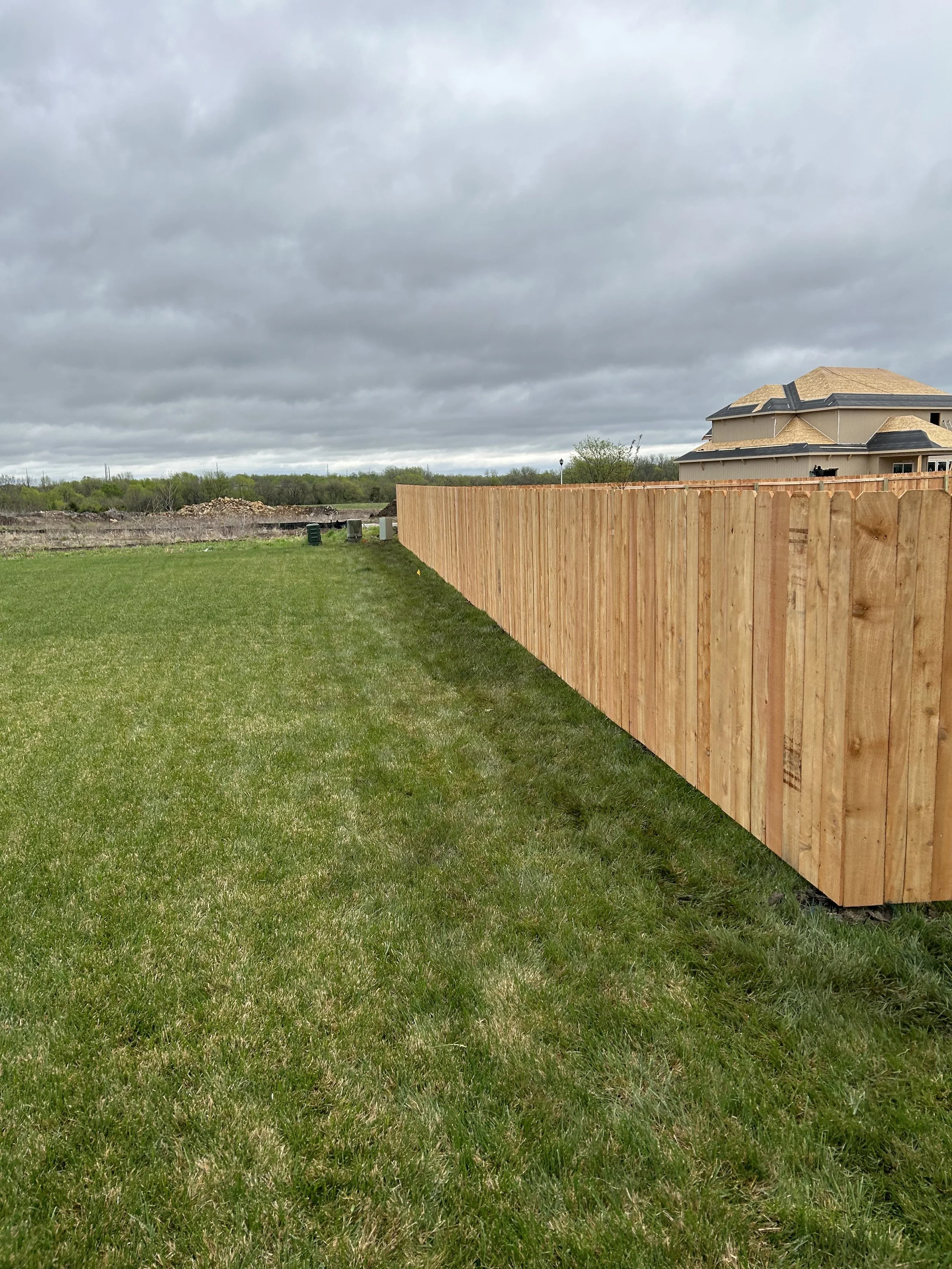 A long wooden privacy fence running parallel to a lush green grassy yard under a cloudy sky with a modern house in the background.