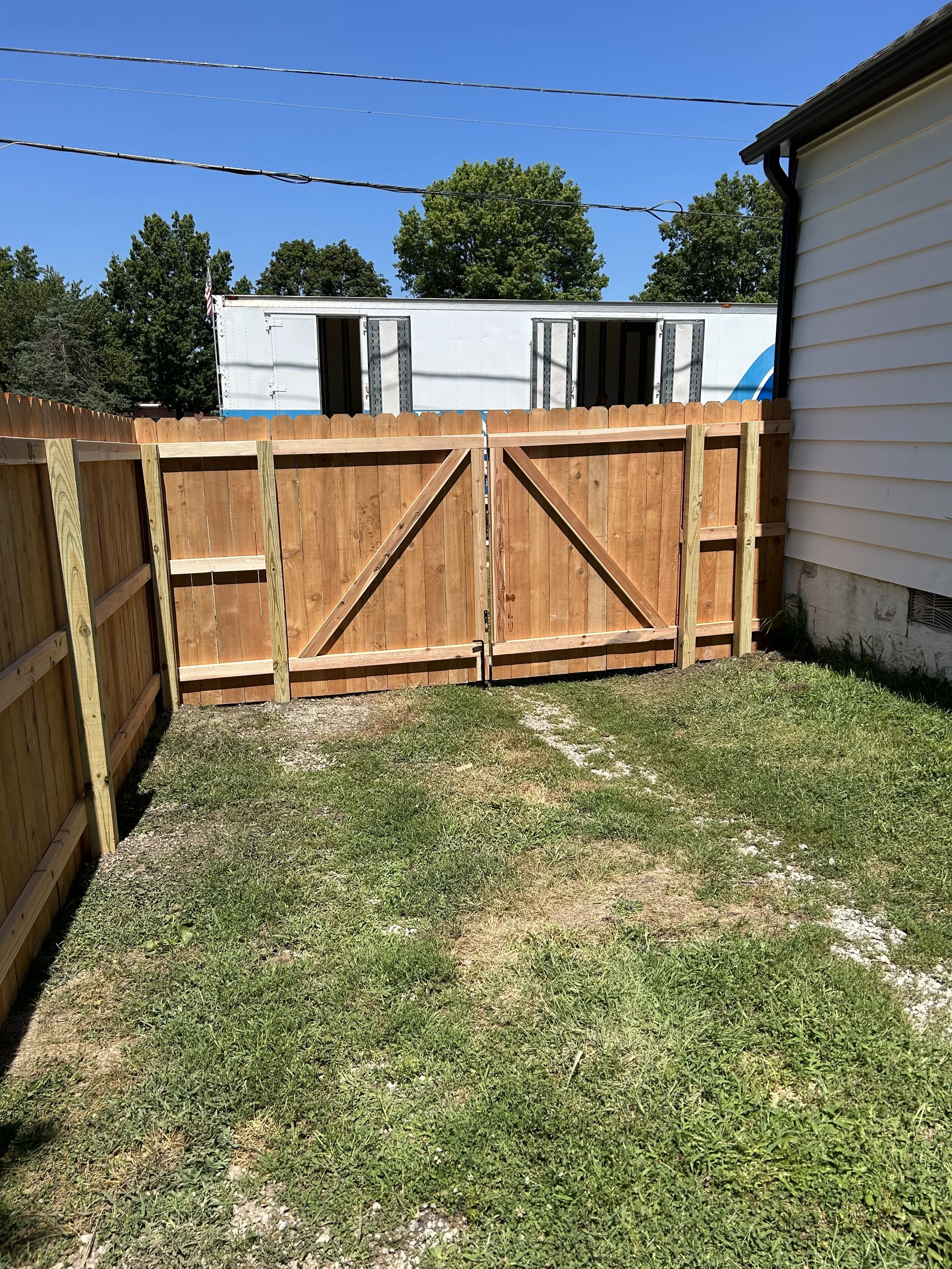 A backyard with a wooden fence and gate, a grassy area, and a white building in the background under a clear blue sky.