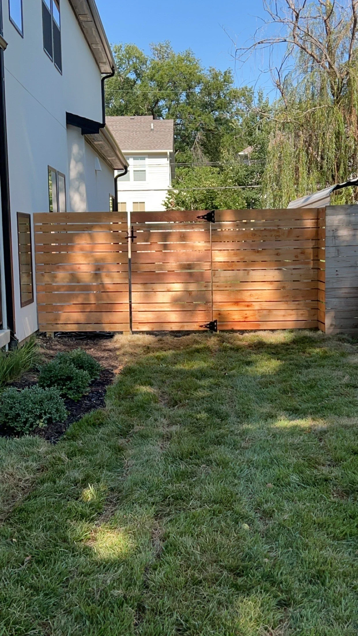 A backyard with a newly installed wooden fence, green grass, small shrubs, and a neighboring house visible in the background under a clear blue sky.