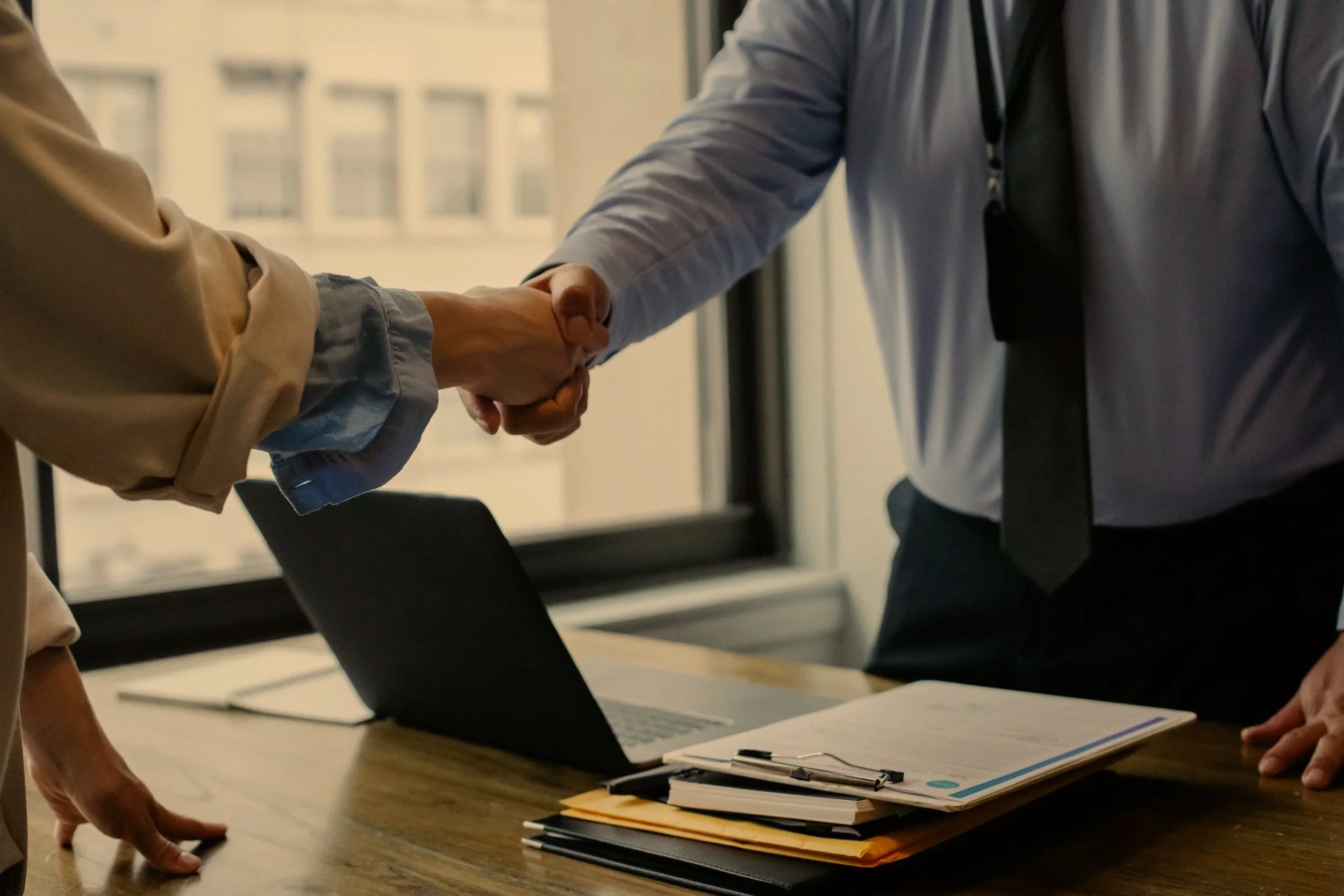 Two people shaking hands in a business setting with a laptop and paperwork on a wooden table in front of them.