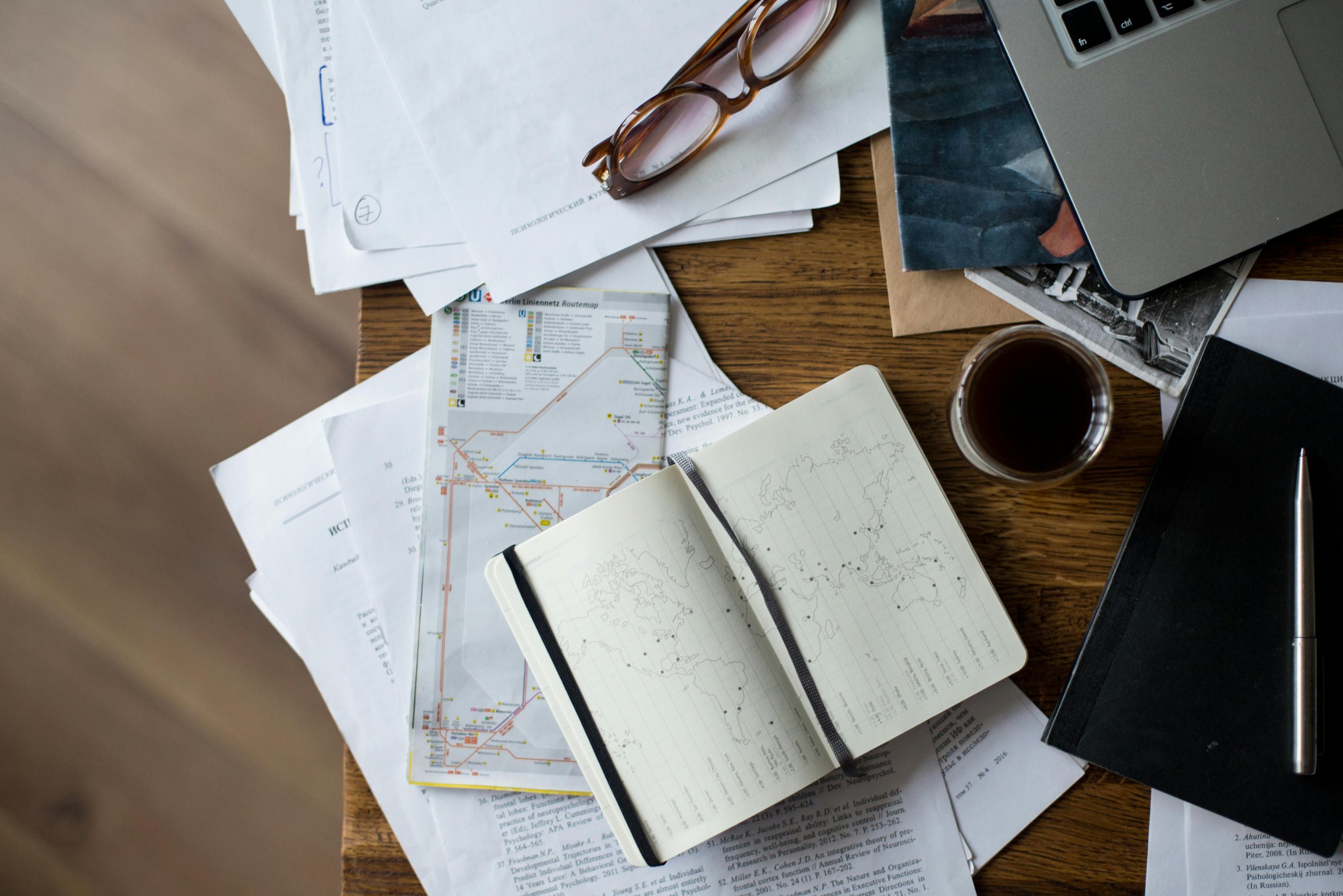 A cluttered wooden desk with scattered papers, maps, a notebook with a world map sketch, a black notebook with a silver pen, a pair of glasses, a laptop, a cup of coffee, and photographs.