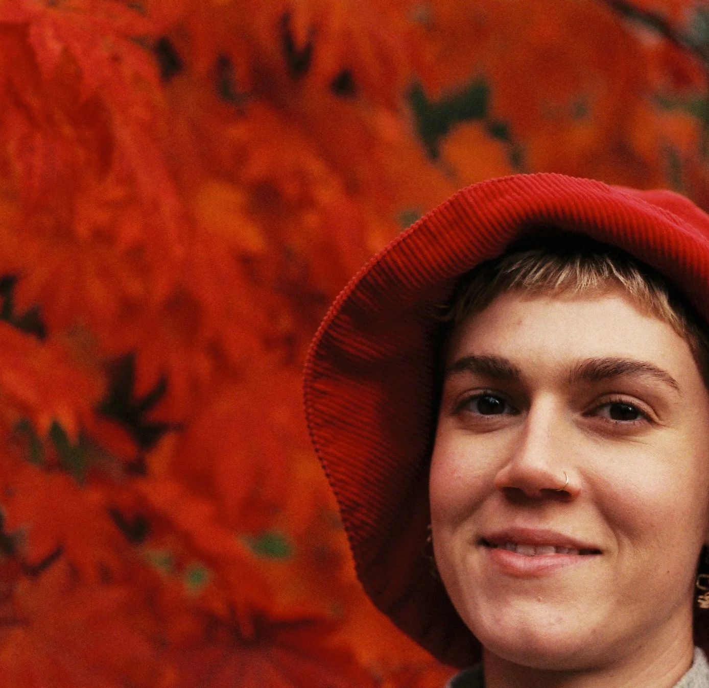 A young person with short hair and a nose ring smiling in front of a vibrant orange and red background of autumn leaves, wearing a wide-brimmed red hat.