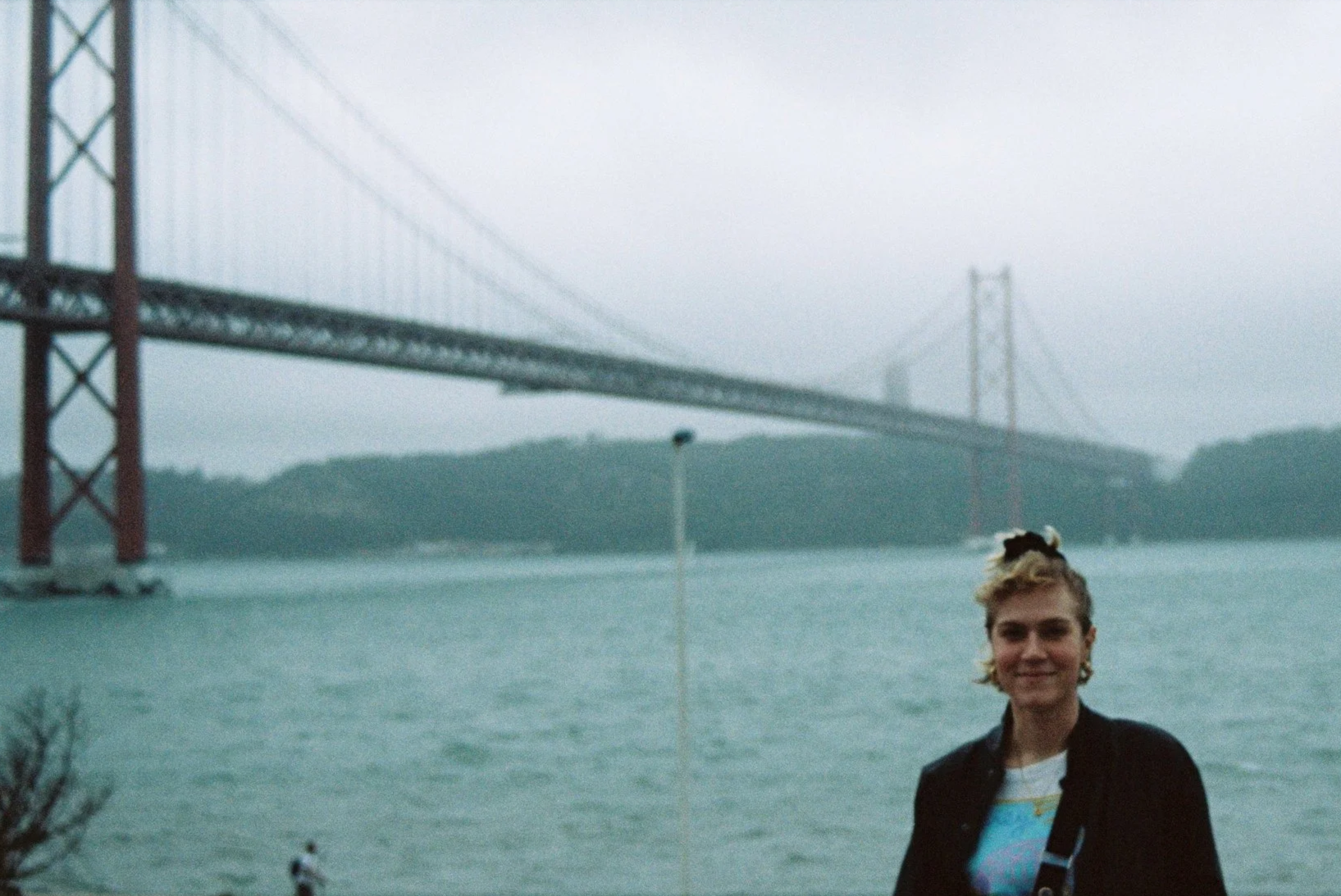 A woman standing near a large body of water with a suspension bridge in the background on a cloudy day.