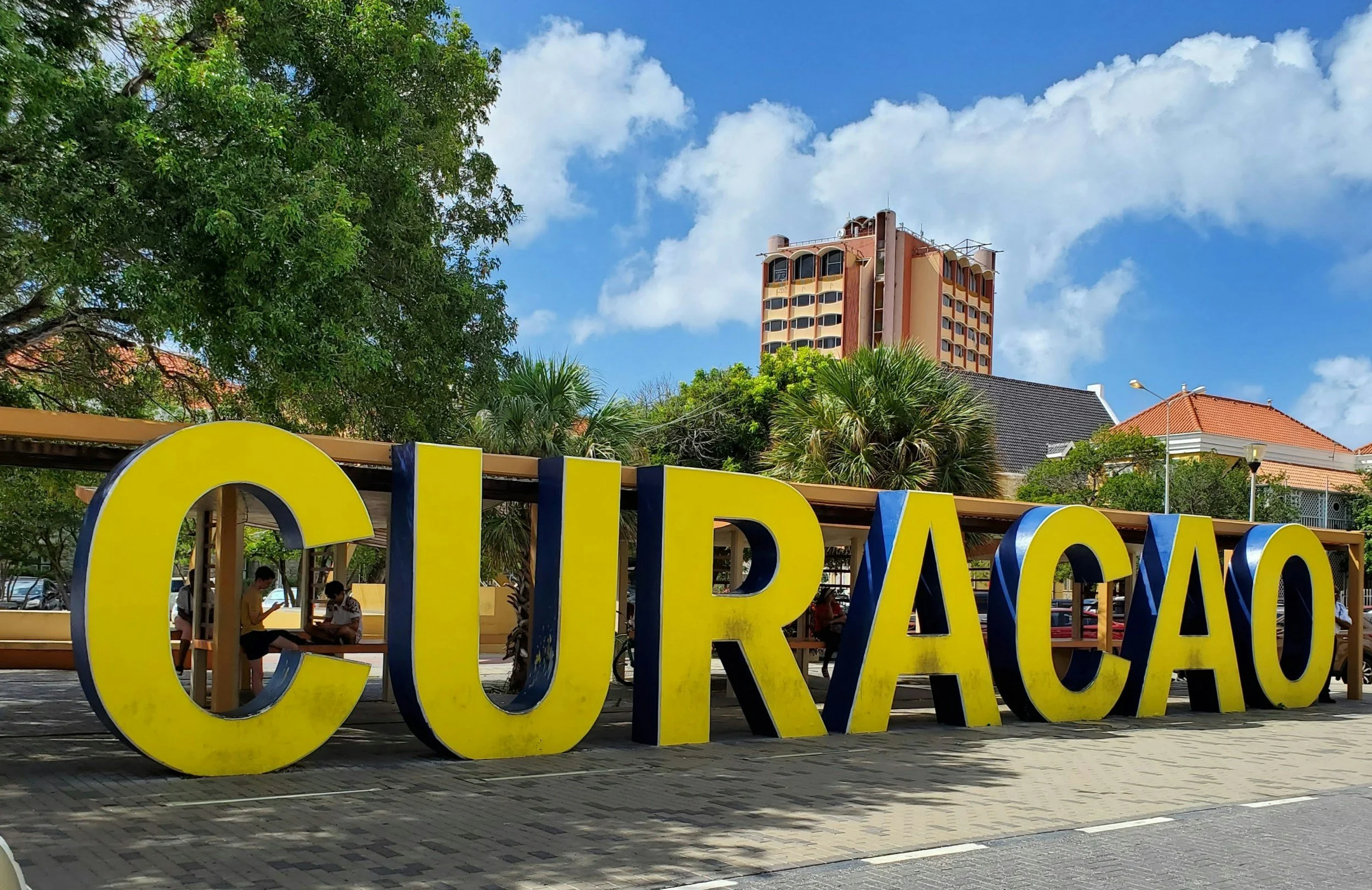 Large yellow and blue 'CURAÇAO' sign outdoors on sidewalk with trees, benches, and buildings in background under partly cloudy sky.