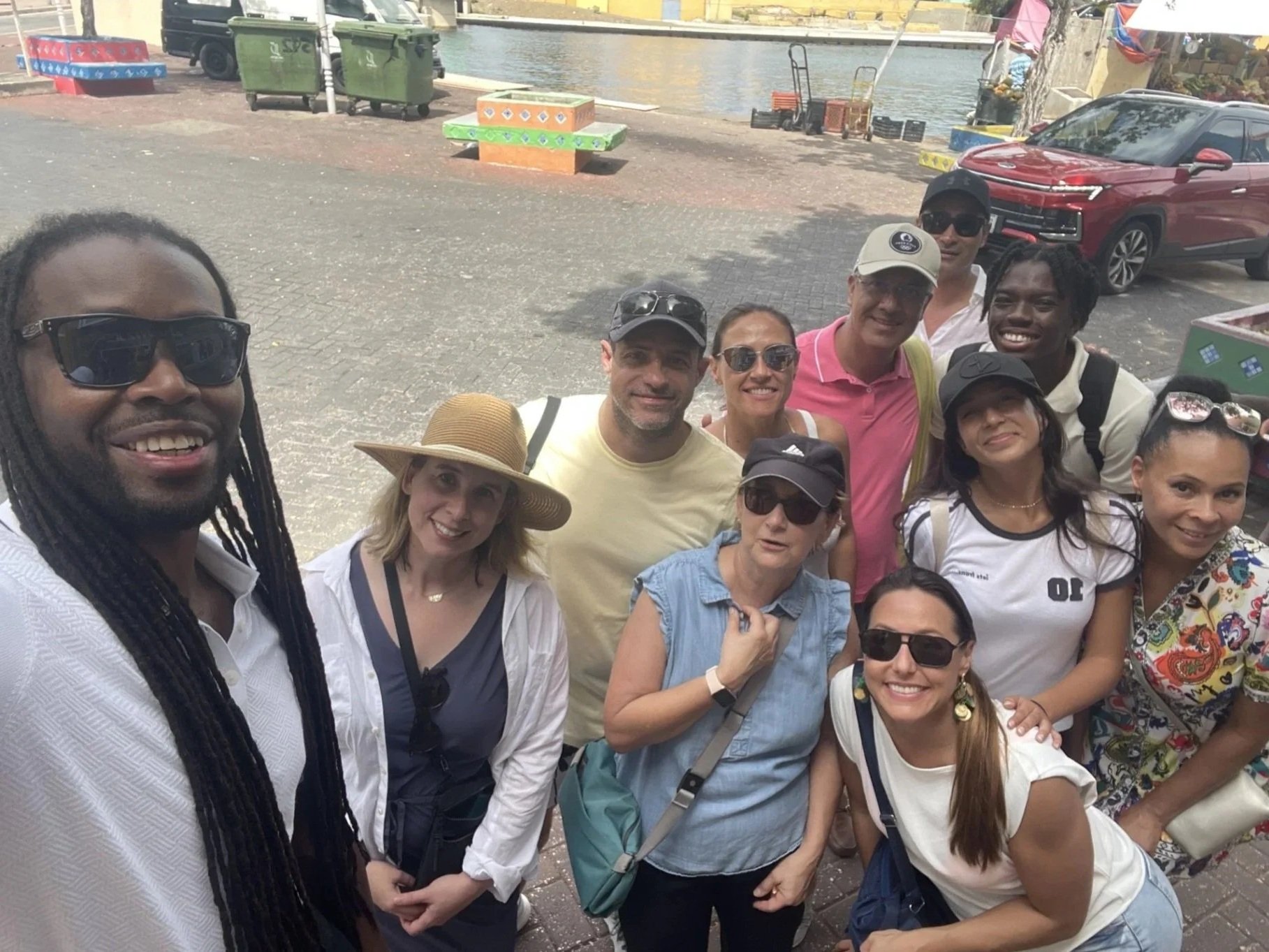 Group of people taking a selfie outdoors near a body of water with colorful benches and parked cars in the background.