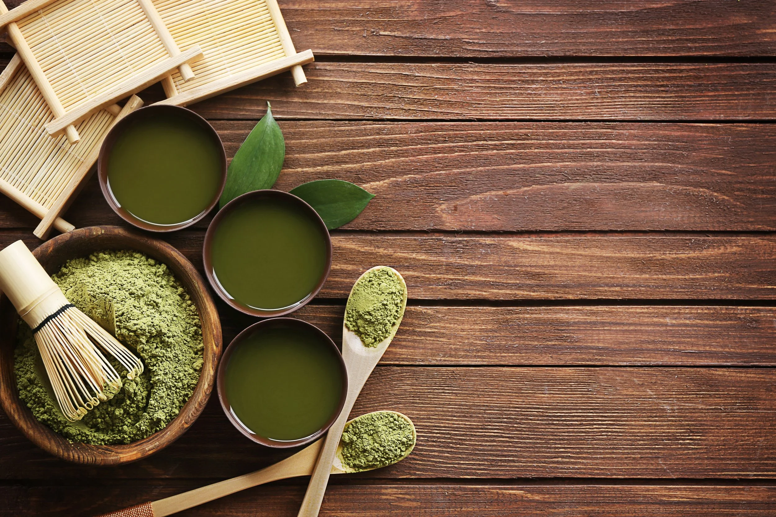 Two cups of matcha tea, matcha powder in a wooden bowl and spoon, bamboo whisk, and bamboo mat on a wooden surface.